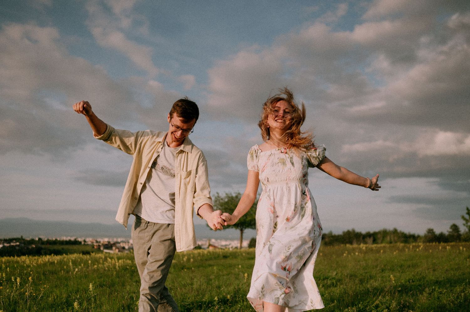 Playful couple dancing and spinning in open field under cloudy sky.