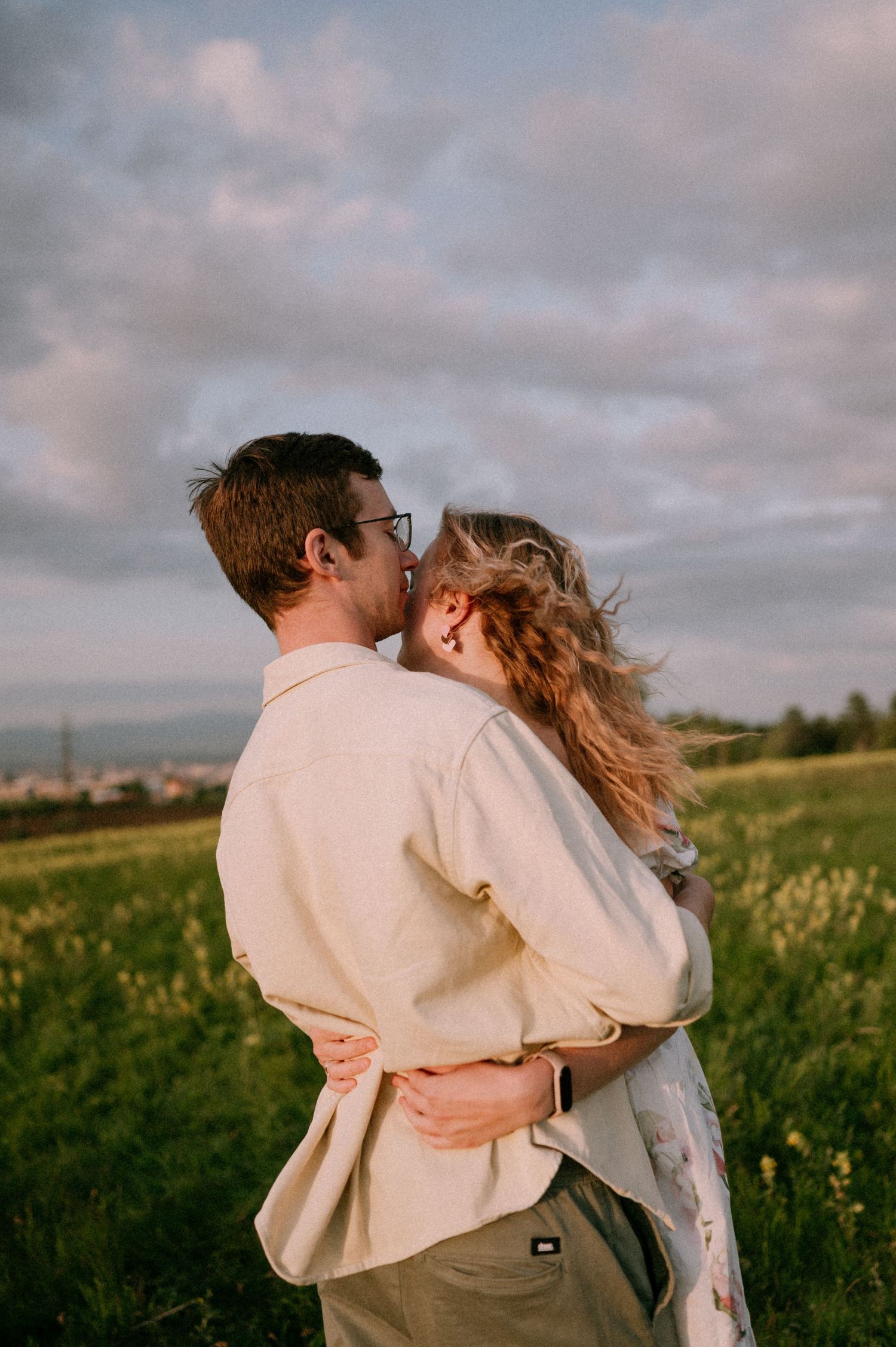 A tender embrace captured in a grassy field during golden hour with cloudy skies overhead.