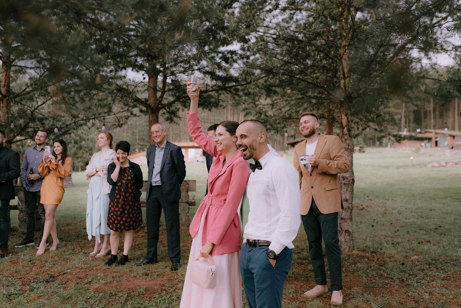 Wedding party and guests gathered for outdoor celebration among pine trees.