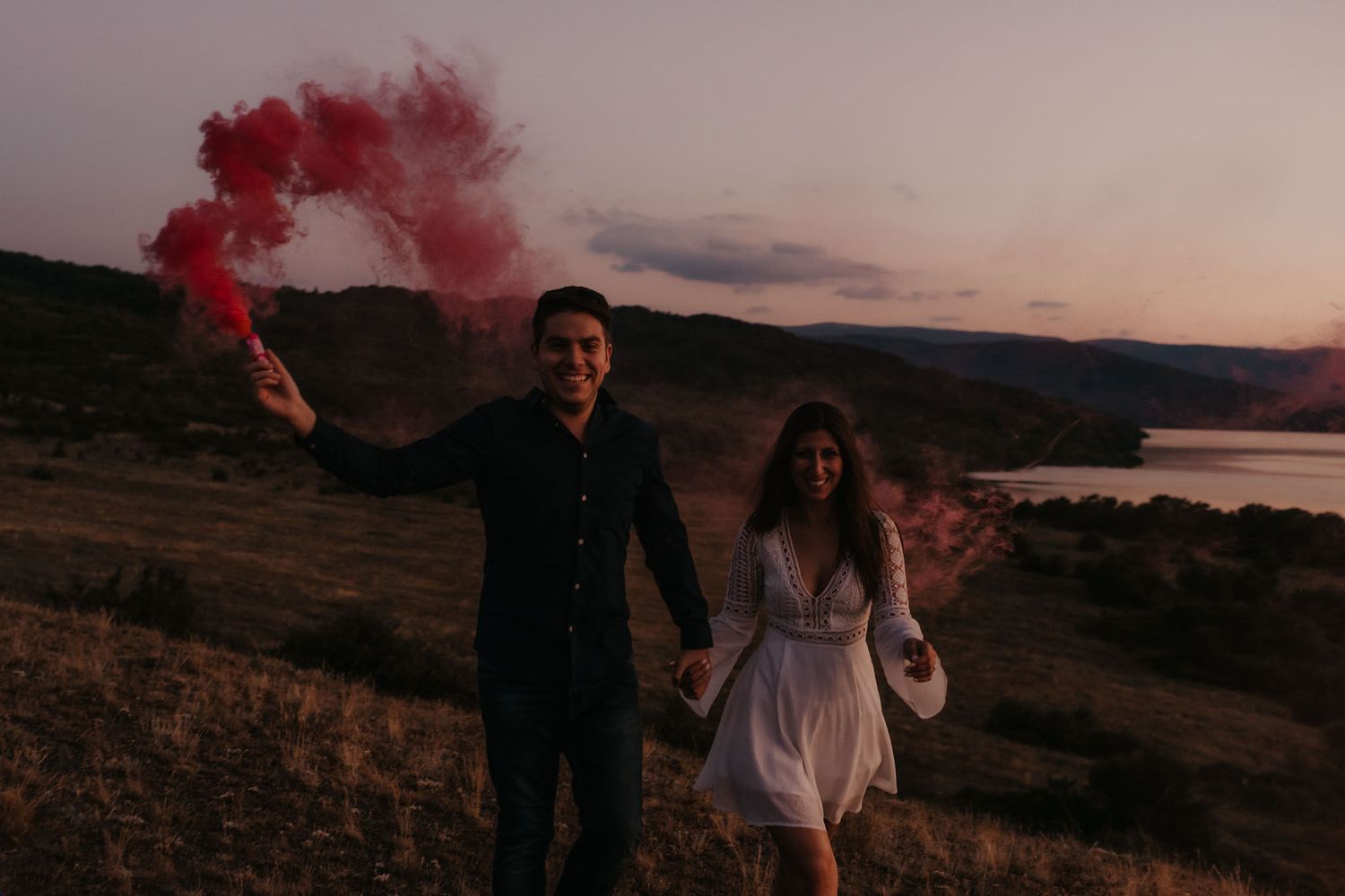 Couple celebrates at sunset on a hilltop with red smoke bombs creating a dramatic atmospheric effect.