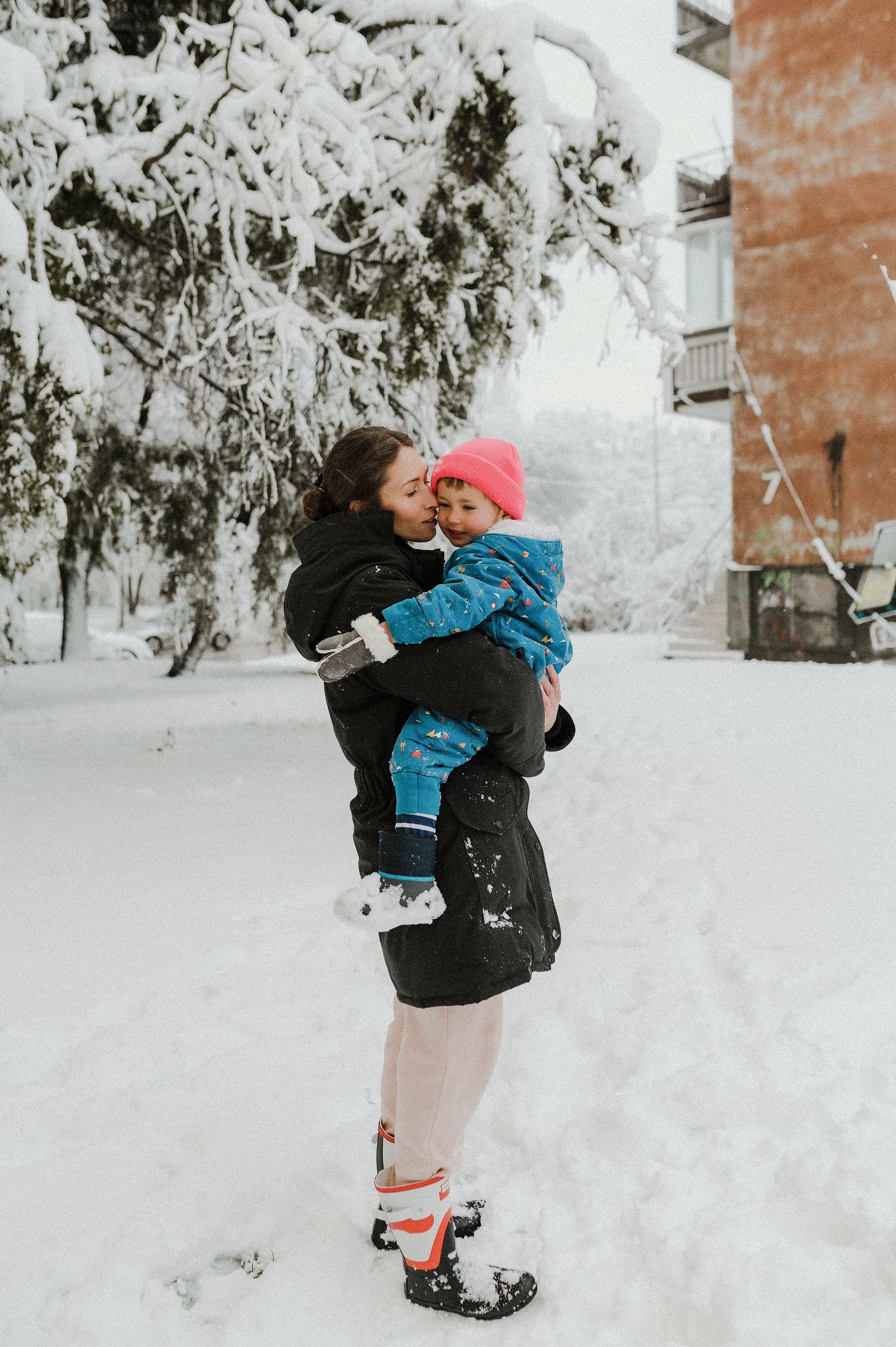 A winter scene shows two people embracing in the snow, with one wearing a bright pink hat and blue snowsuit.