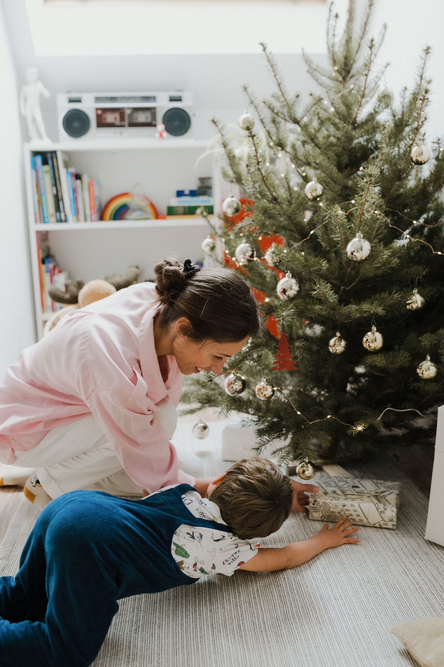 Two people sit on the floor beside a decorated Christmas tree with ornaments and lights in a cozy living room setting.