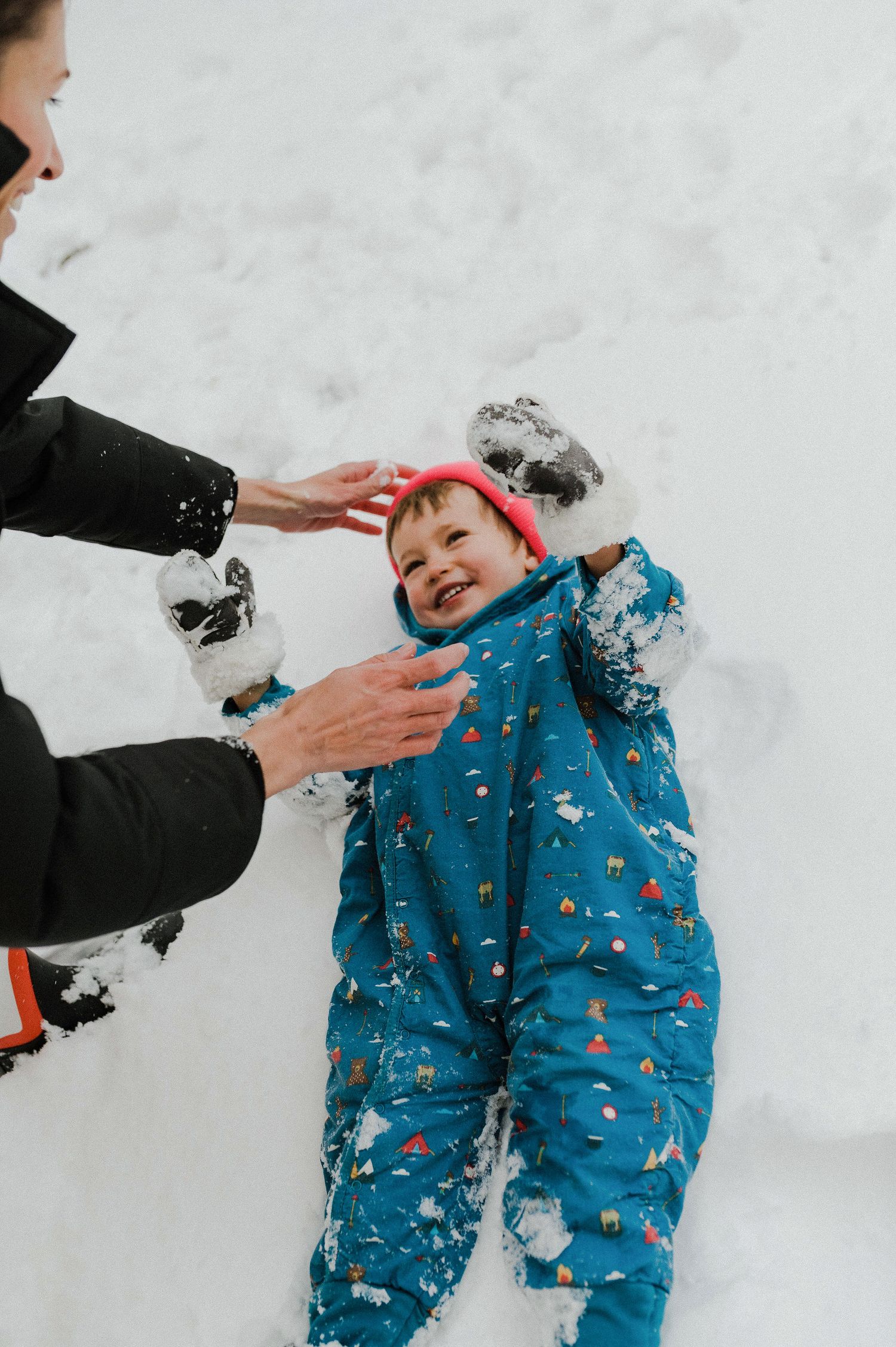 Child wearing blue snowsuit with pattern plays in fresh snow while hands reach down to help them play.