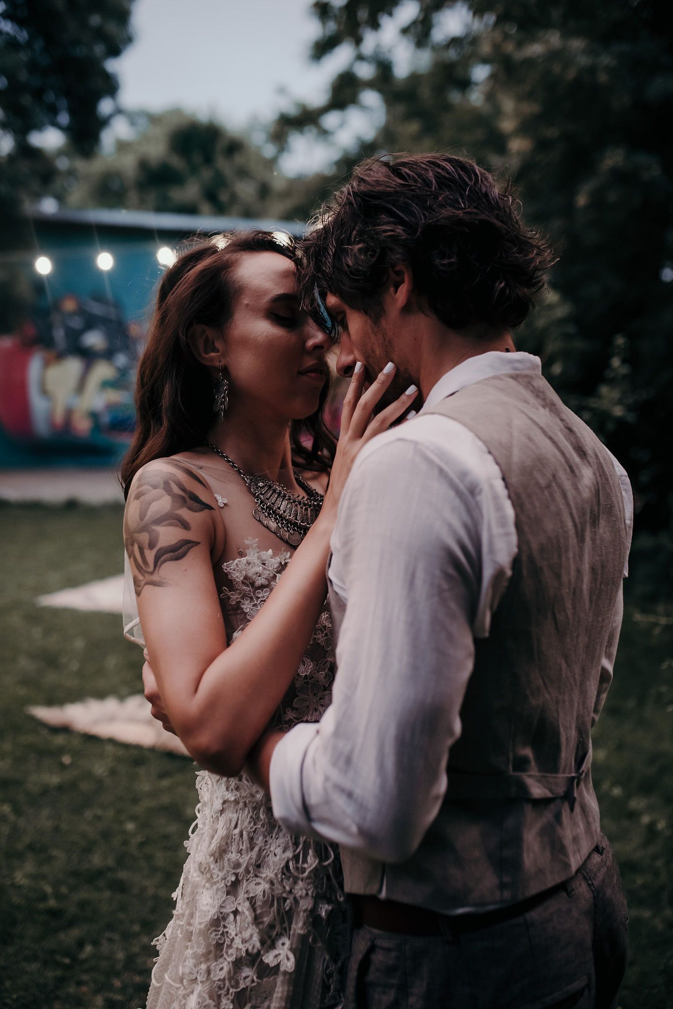 A couple in vintage-style wedding attire share a romantic moment outdoors in a park setting.