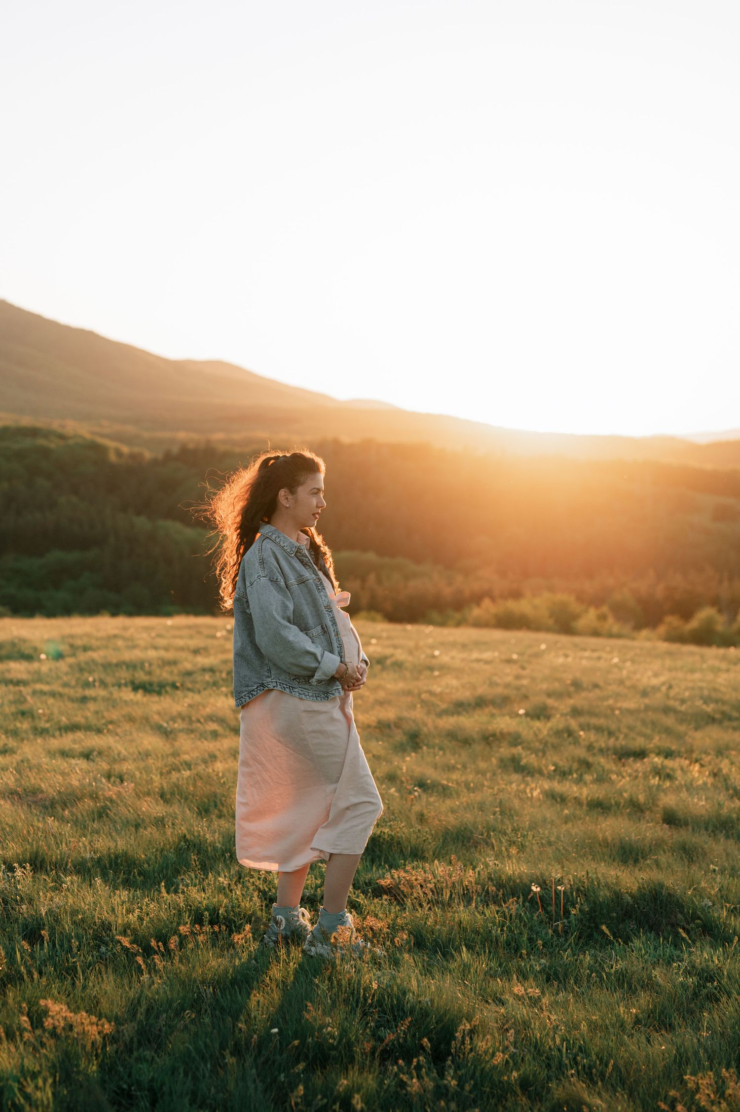 Person in denim jacket and skirt stands in grassy field during golden sunset hour.