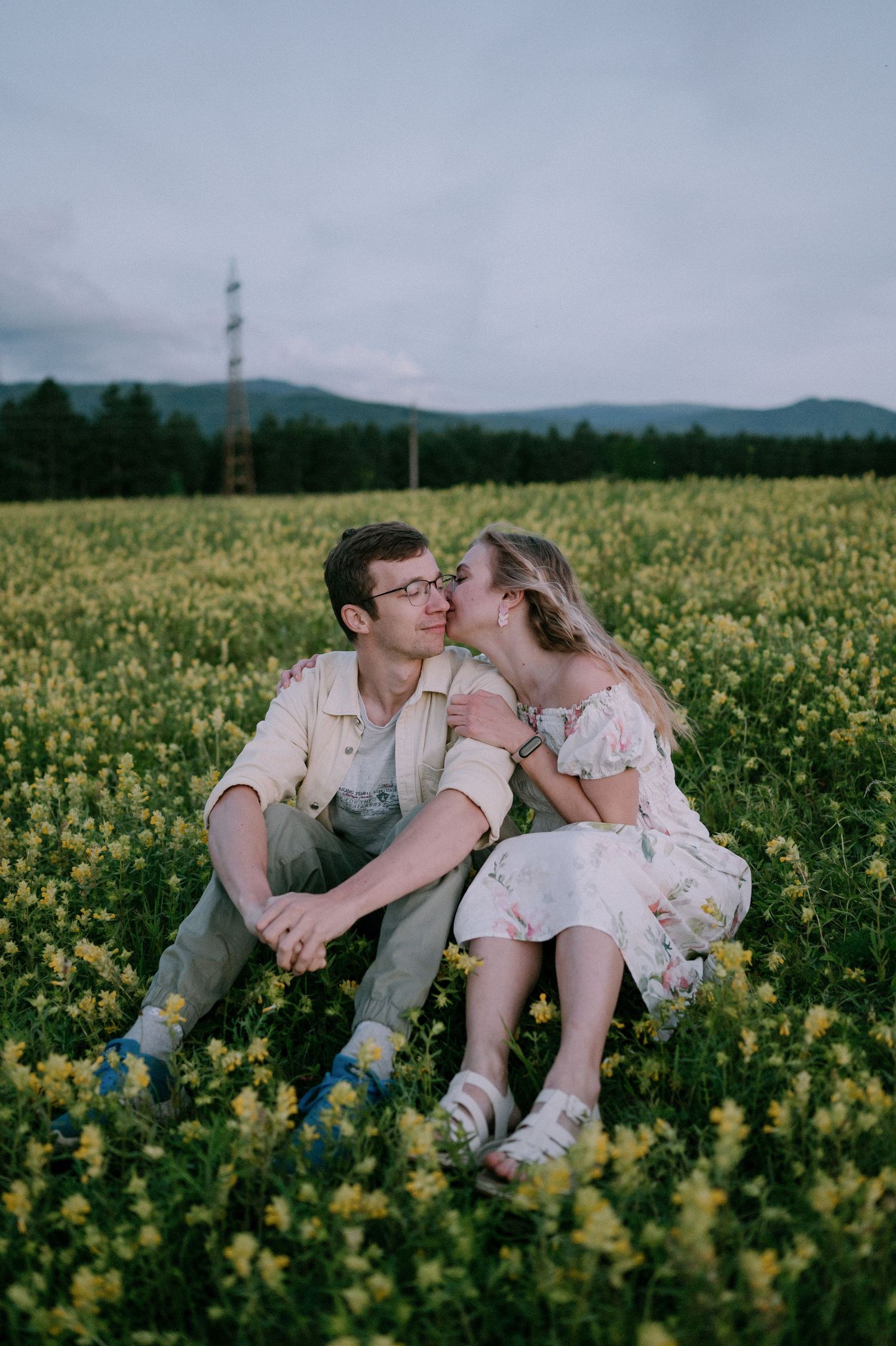 Two people share an intimate kiss while sitting in a field of yellow wildflowers.