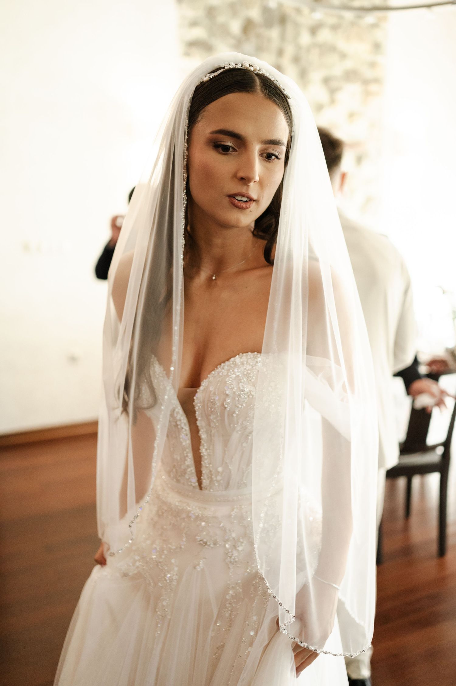 A bride in an elegant white lace gown and flowing veil poses in a bright indoor setting.