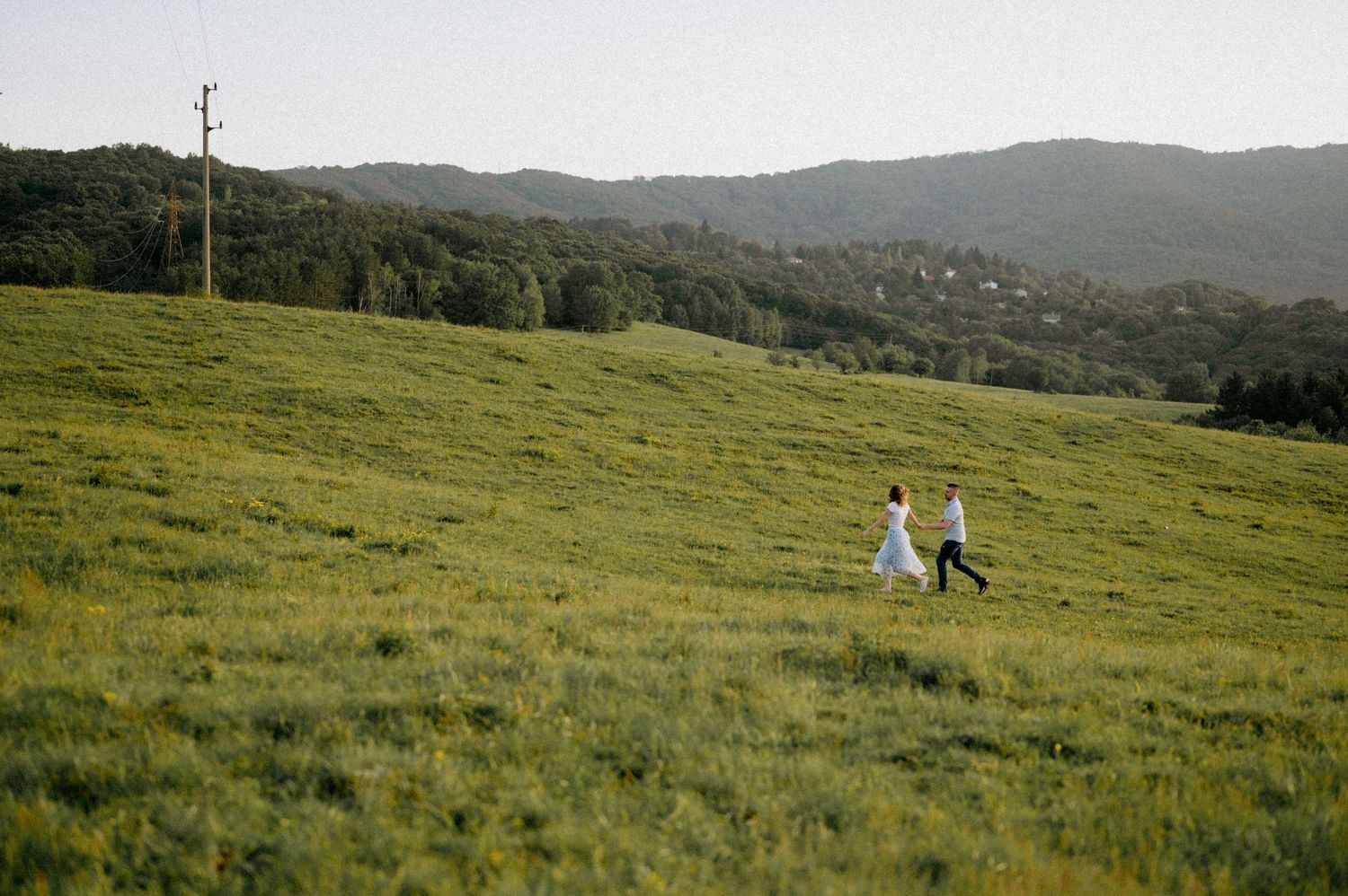 Two people run playfully across a grassy field with mountains and utility poles in the background.