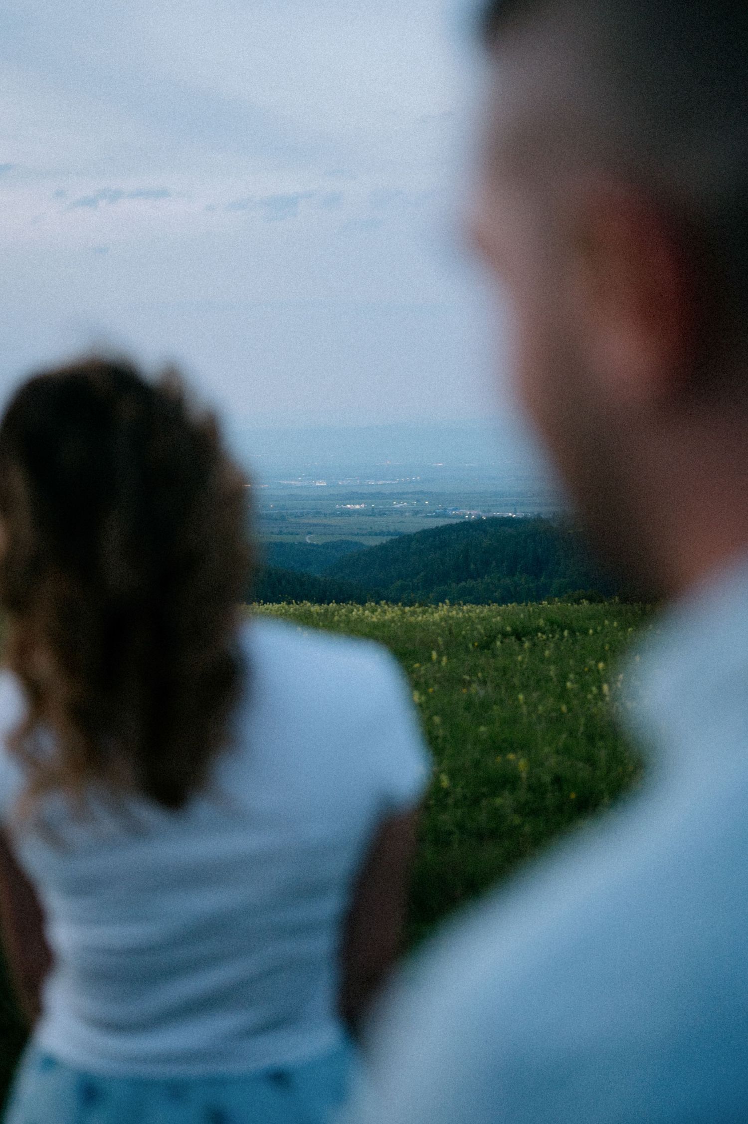 A couple stands together on a hilltop overlooking a scenic landscape at dusk.