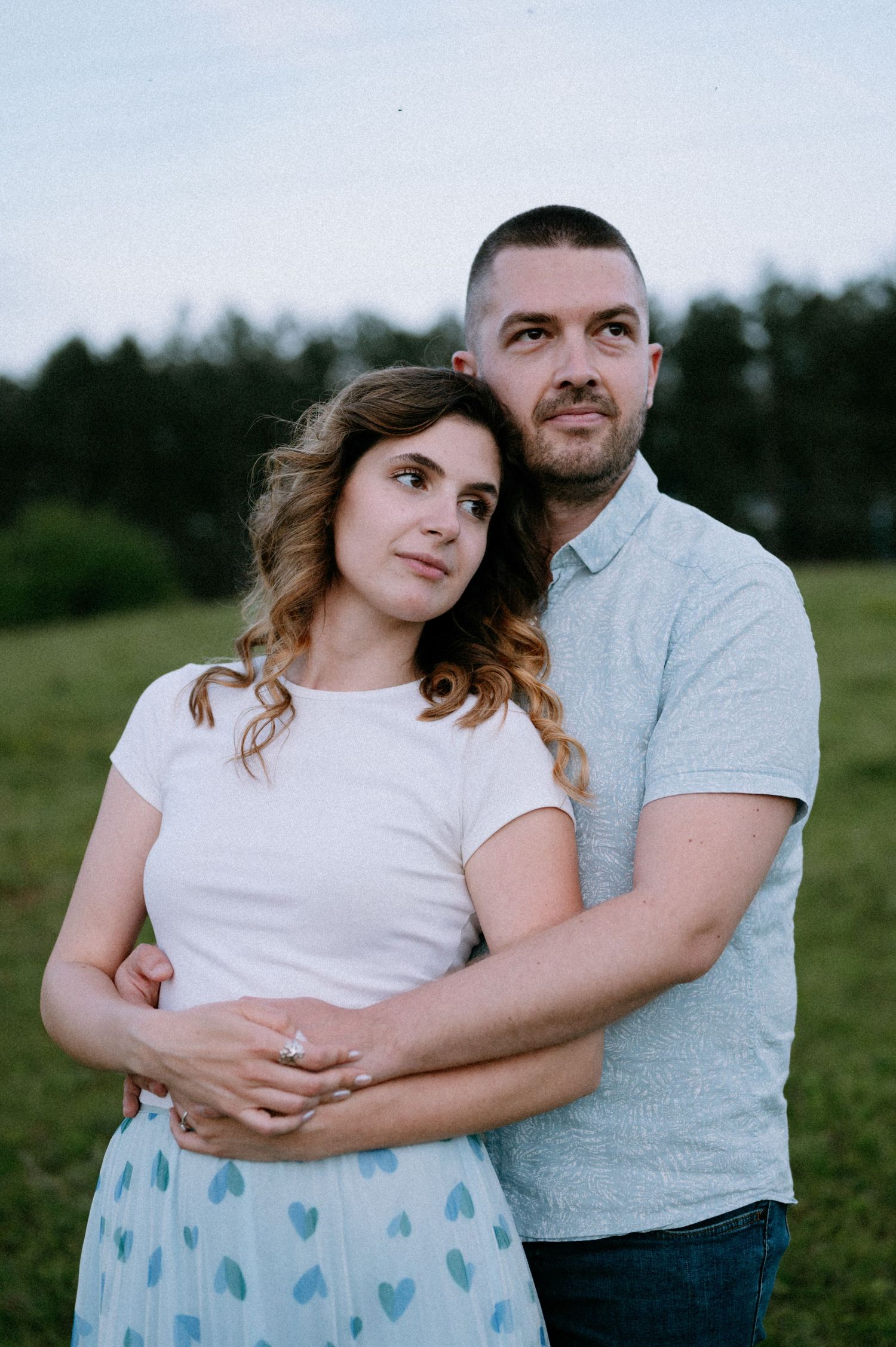 A couple embraces in a grassy field wearing casual clothing during an outdoor photoshoot.