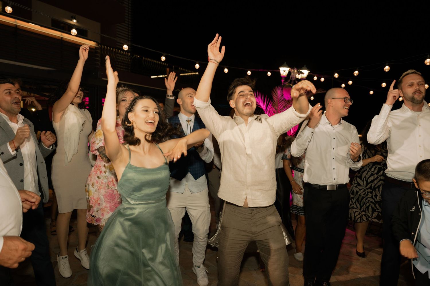 Wedding guests dance and celebrate with raised arms on a dimly lit dance floor during a lively reception party.