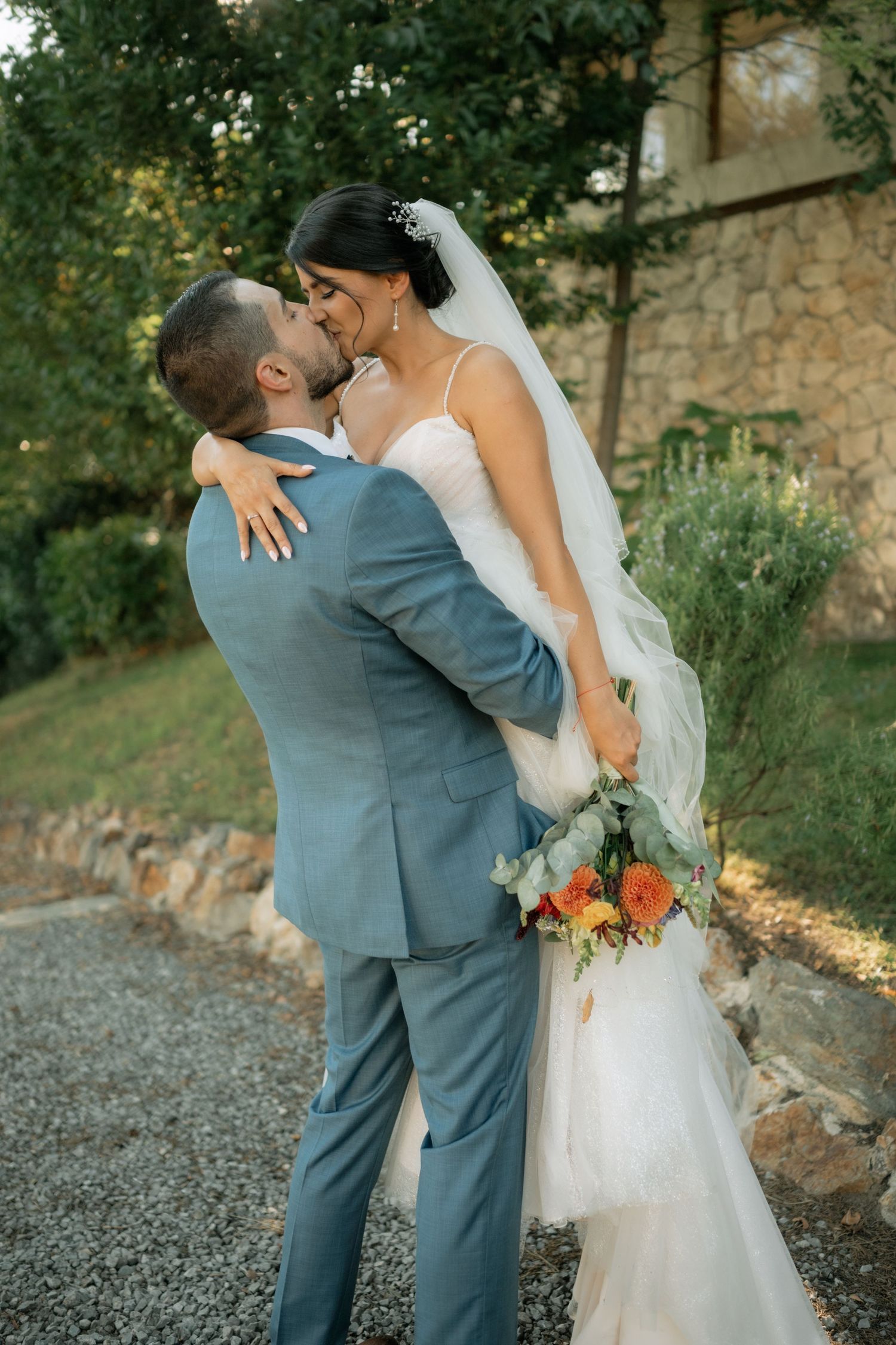 A romantic wedding couple embraces outdoors with the bride holding a vibrant orange and green bouquet.
