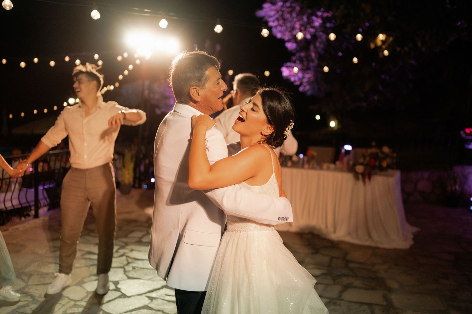 Wedding guests dance and celebrate on an outdoor dance floor illuminated by string lights at night.