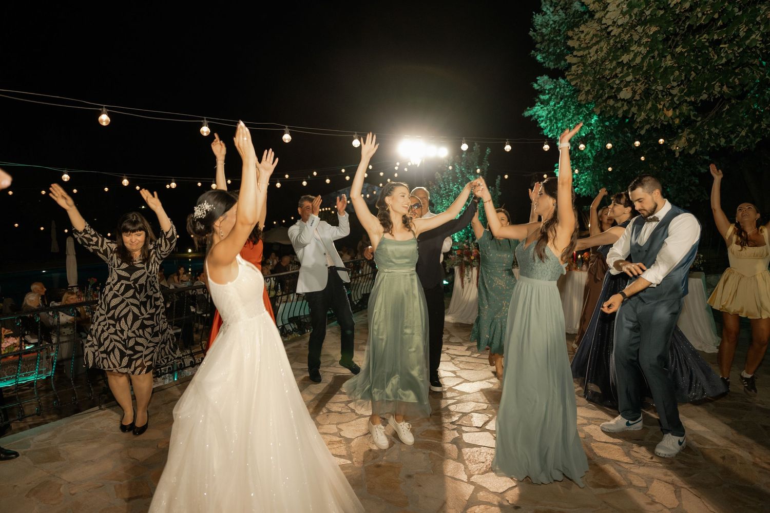Wedding guests dancing together outdoors at night under string lights during a festive reception celebration.