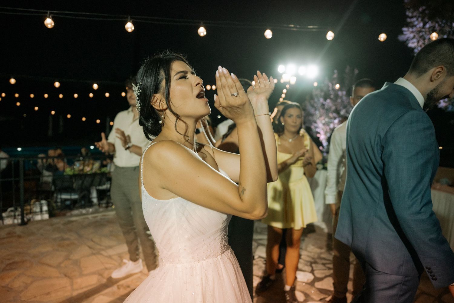 Wedding guests dance and celebrate on an outdoor dance floor illuminated by string lights and spotlights at night.