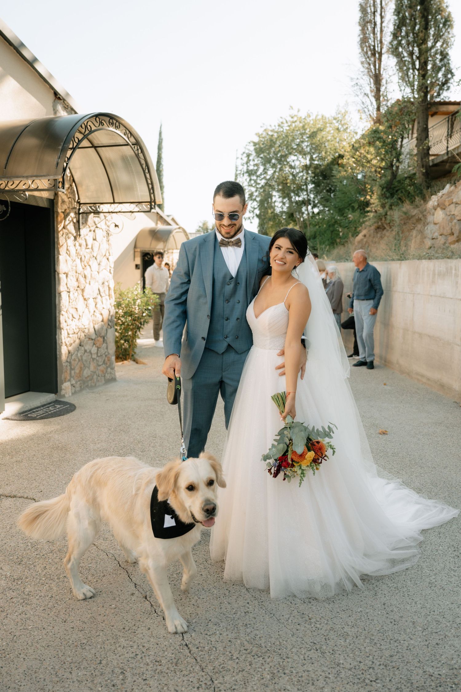 A couple in wedding attire walks down a stone path with their golden retriever dog following behind them.