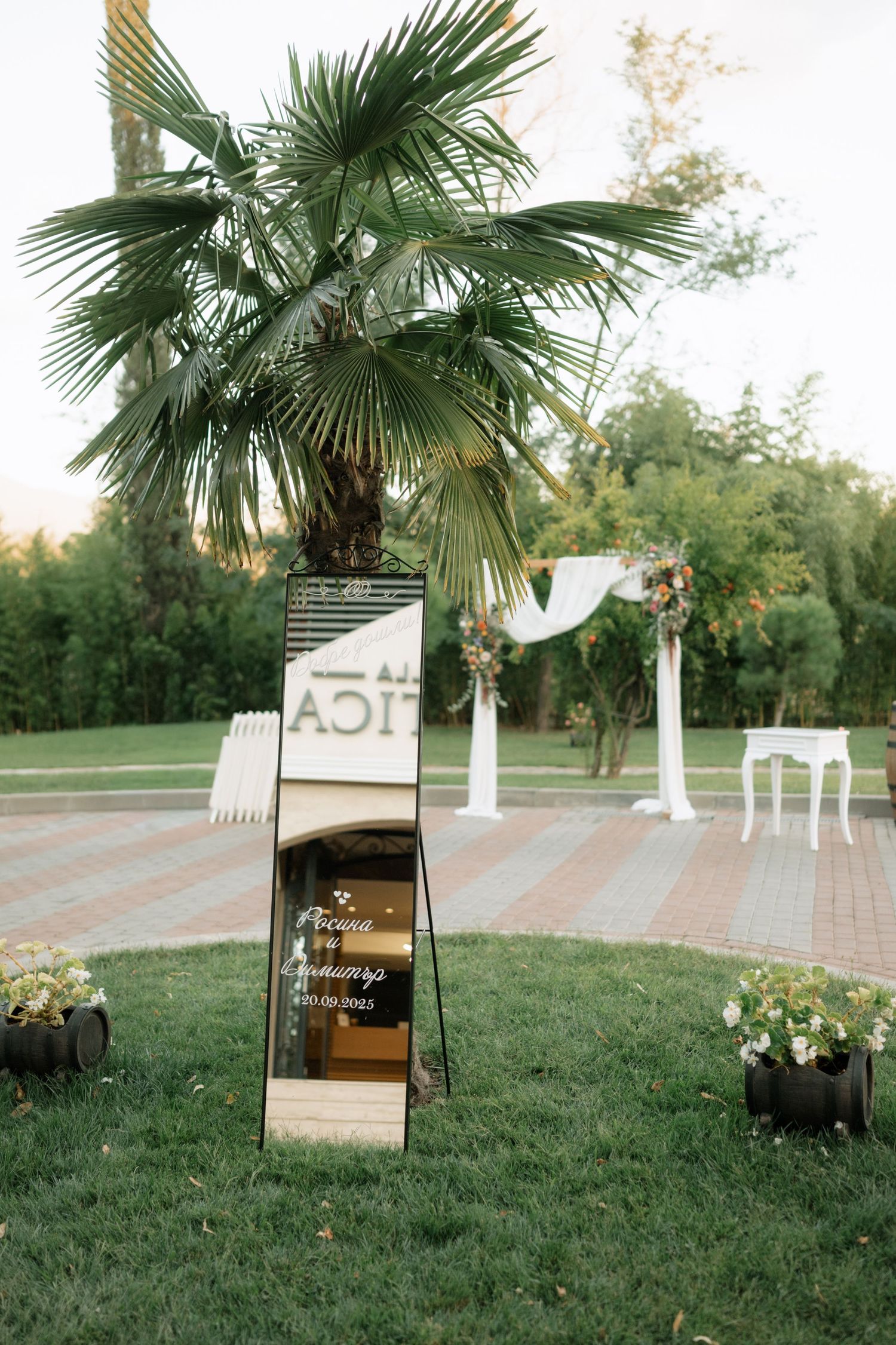 Outdoor wedding ceremony setup with a mirrored sign, white arch, and palm tree in an elegant garden setting.
