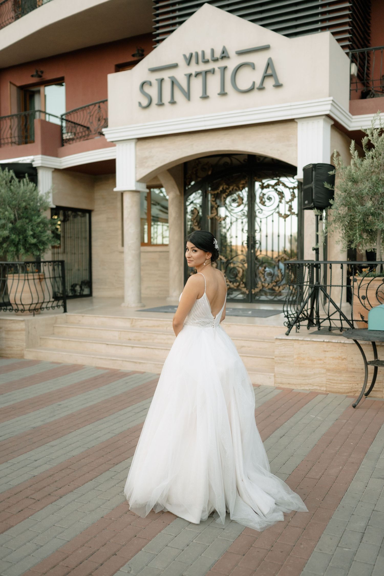 A bride in a flowing white wedding dress stands in front of Villa Sintica's entrance during a special occasion.