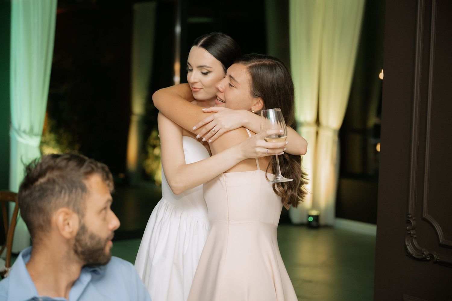 Two people share a warm embrace at an elegant indoor event with soft green drapery in the background.