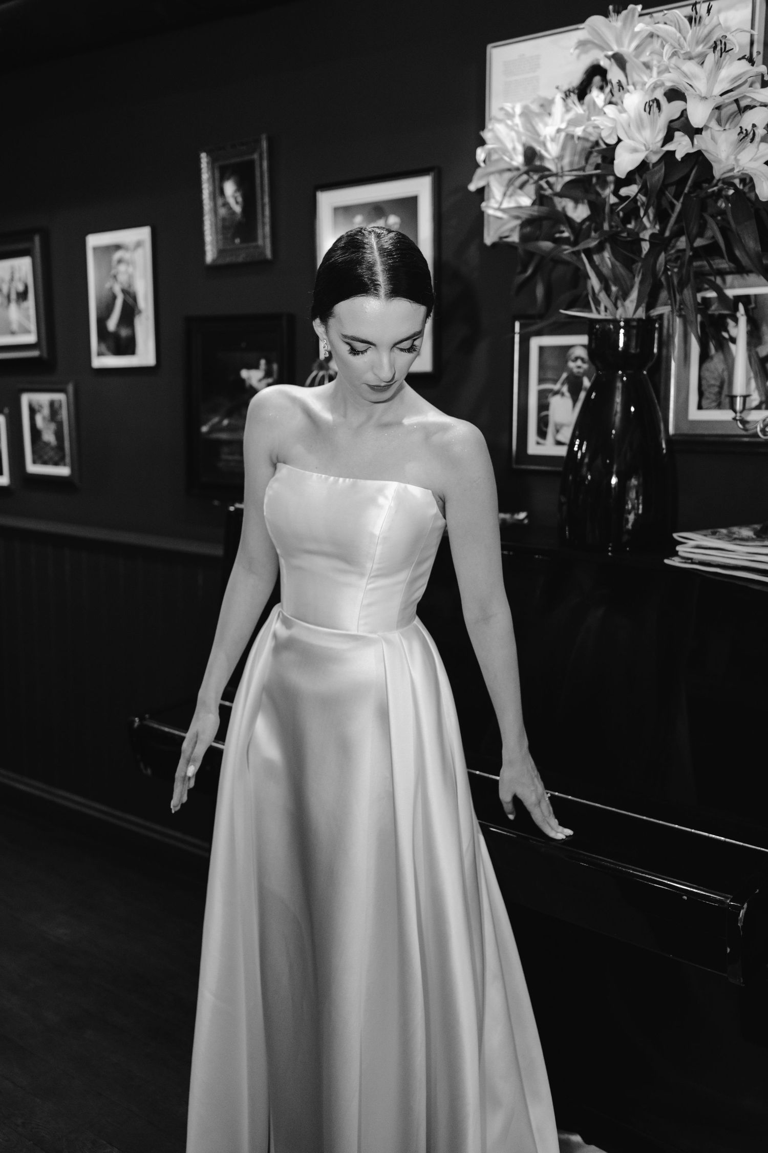 A person wearing a strapless white formal gown stands in a dark room with framed photos on the wall.