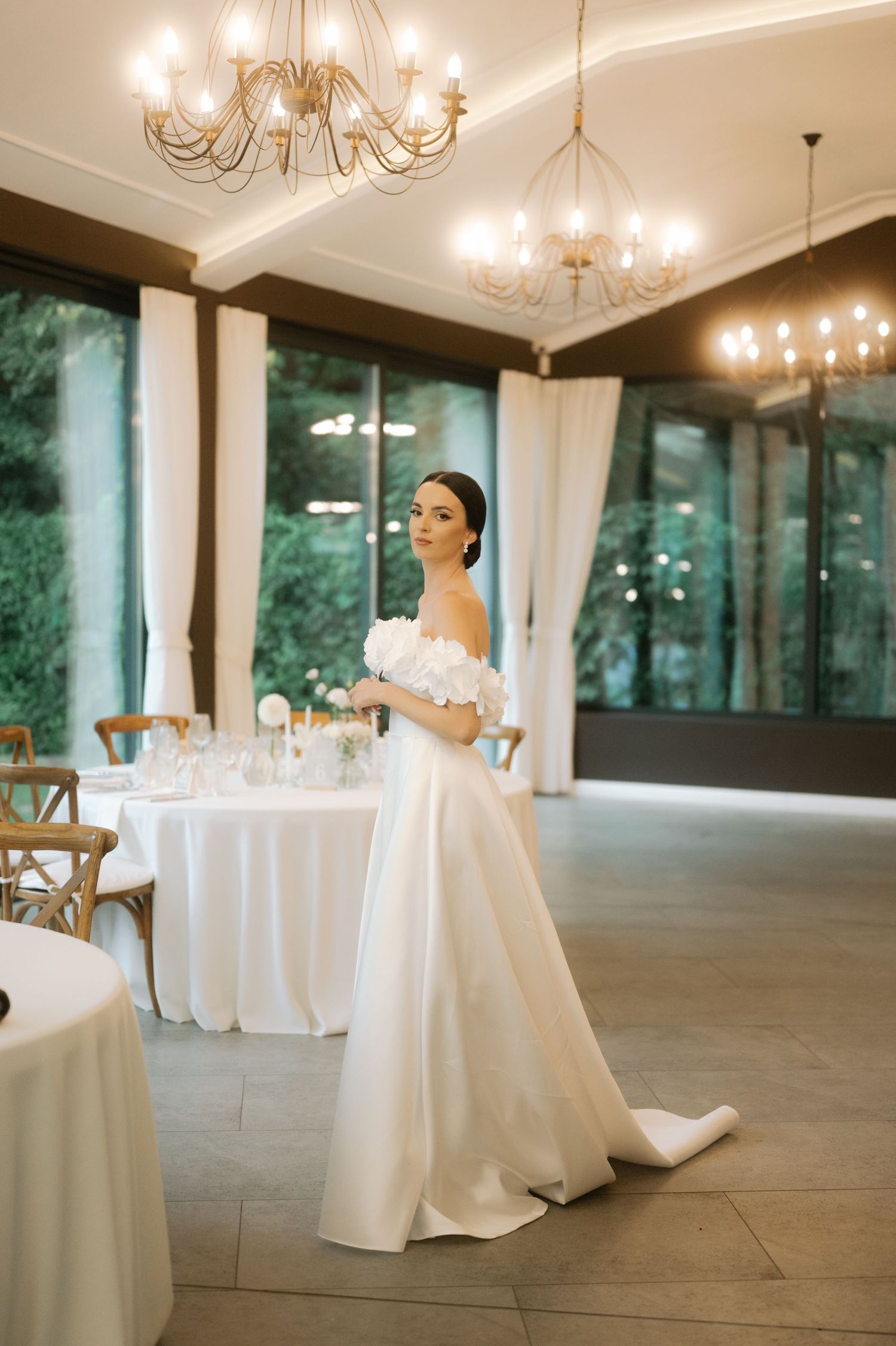 Elegant wedding reception venue with white tablecloths and chandeliers in a spacious room with large windows.