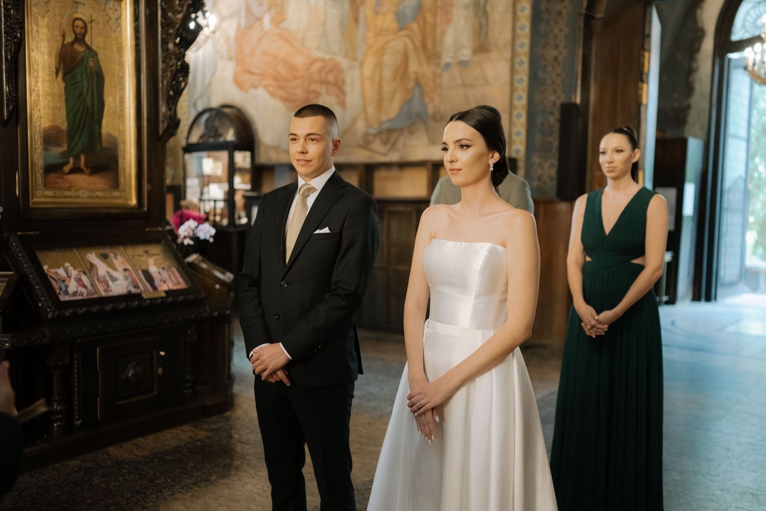 Wedding ceremony participants stand in a church with ornate religious artwork and icons on the wall behind them.