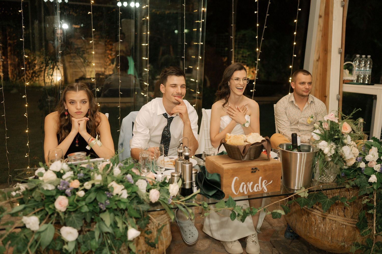 Wedding party members laugh and react emotionally while seated at a rustic reception table decorated with greenery garlands.