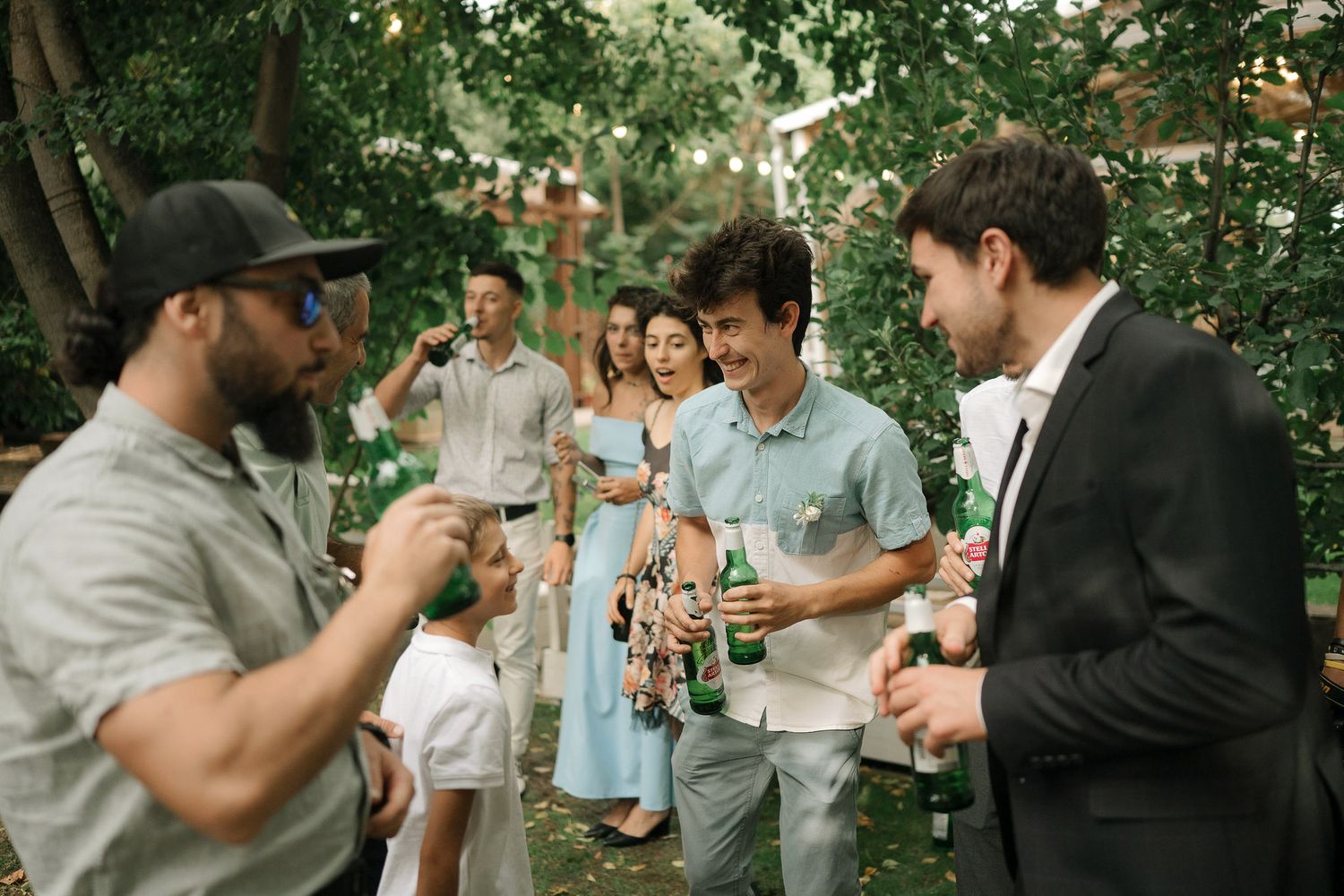 A casual outdoor gathering with people holding drinks and socializing under green trees on a sunny day.