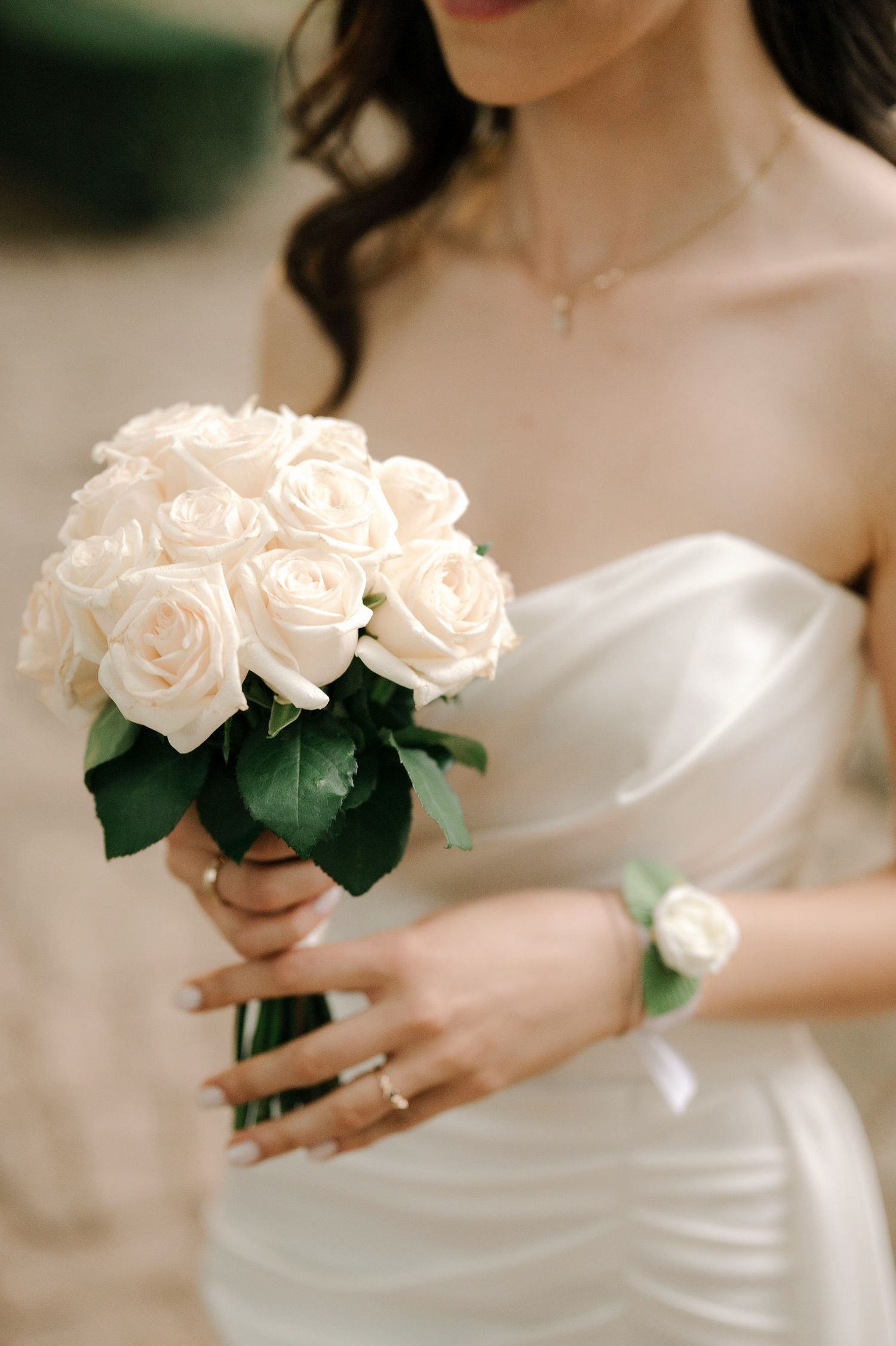 A delicate white rose wedding bouquet held by a bride in a white dress with matching floral wrist corsage.