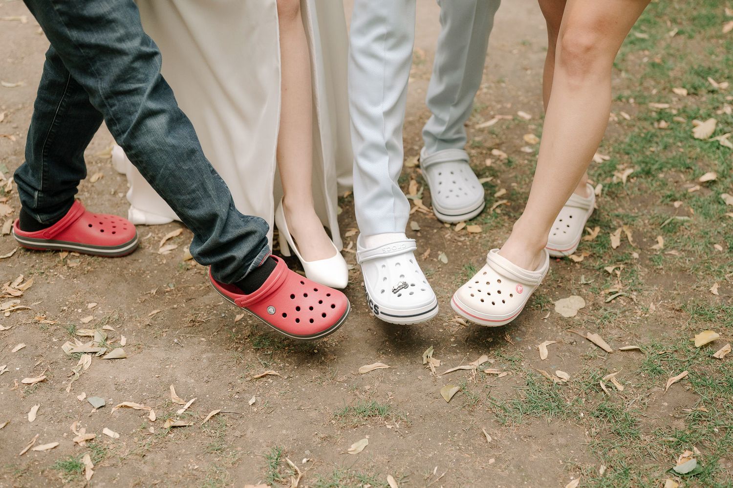 A row of feet wearing various styles of Crocs shoes stand together on a dirt path.