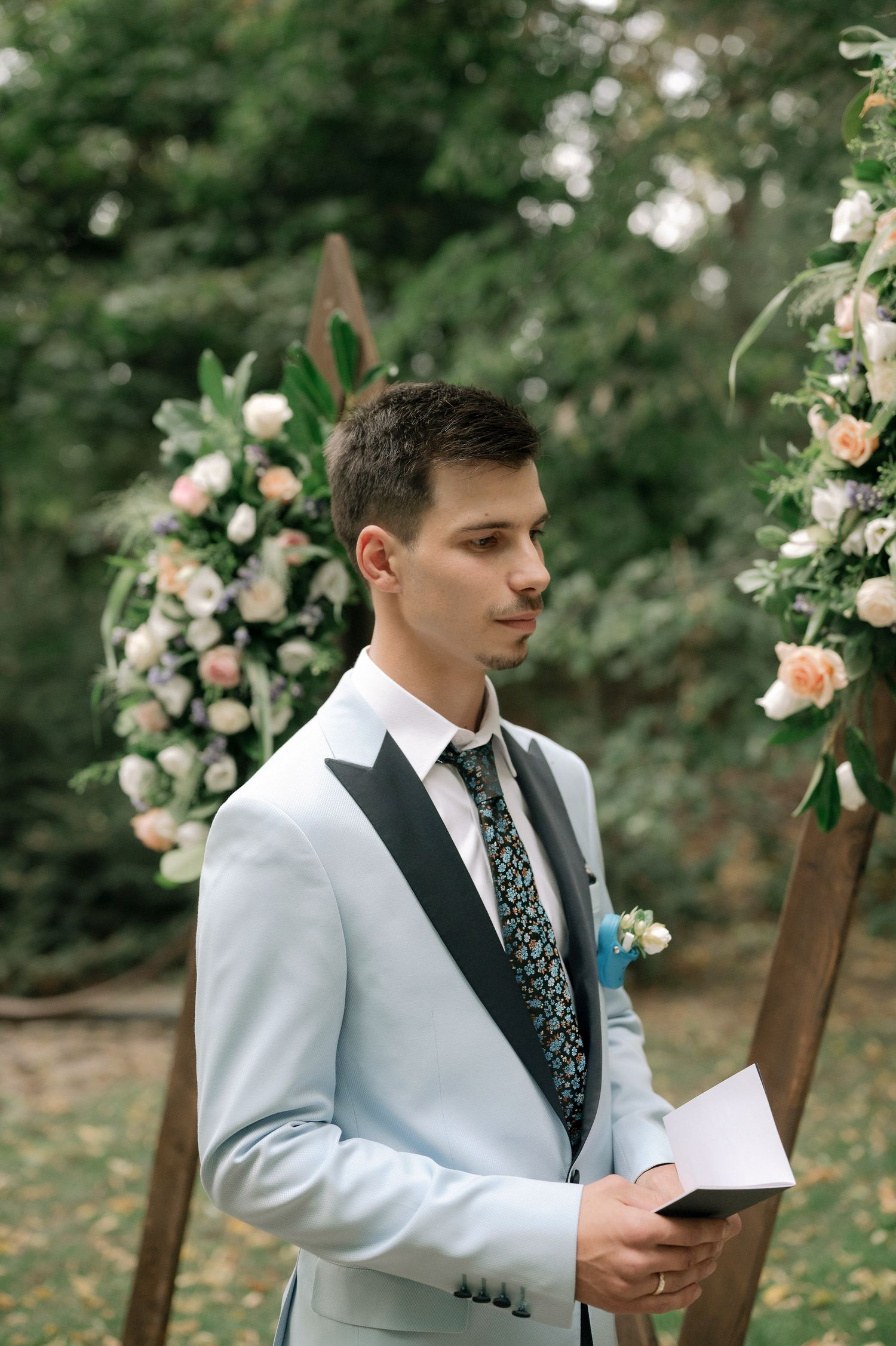 A groom in a white suit and patterned tie reads from a book during an outdoor garden wedding ceremony.