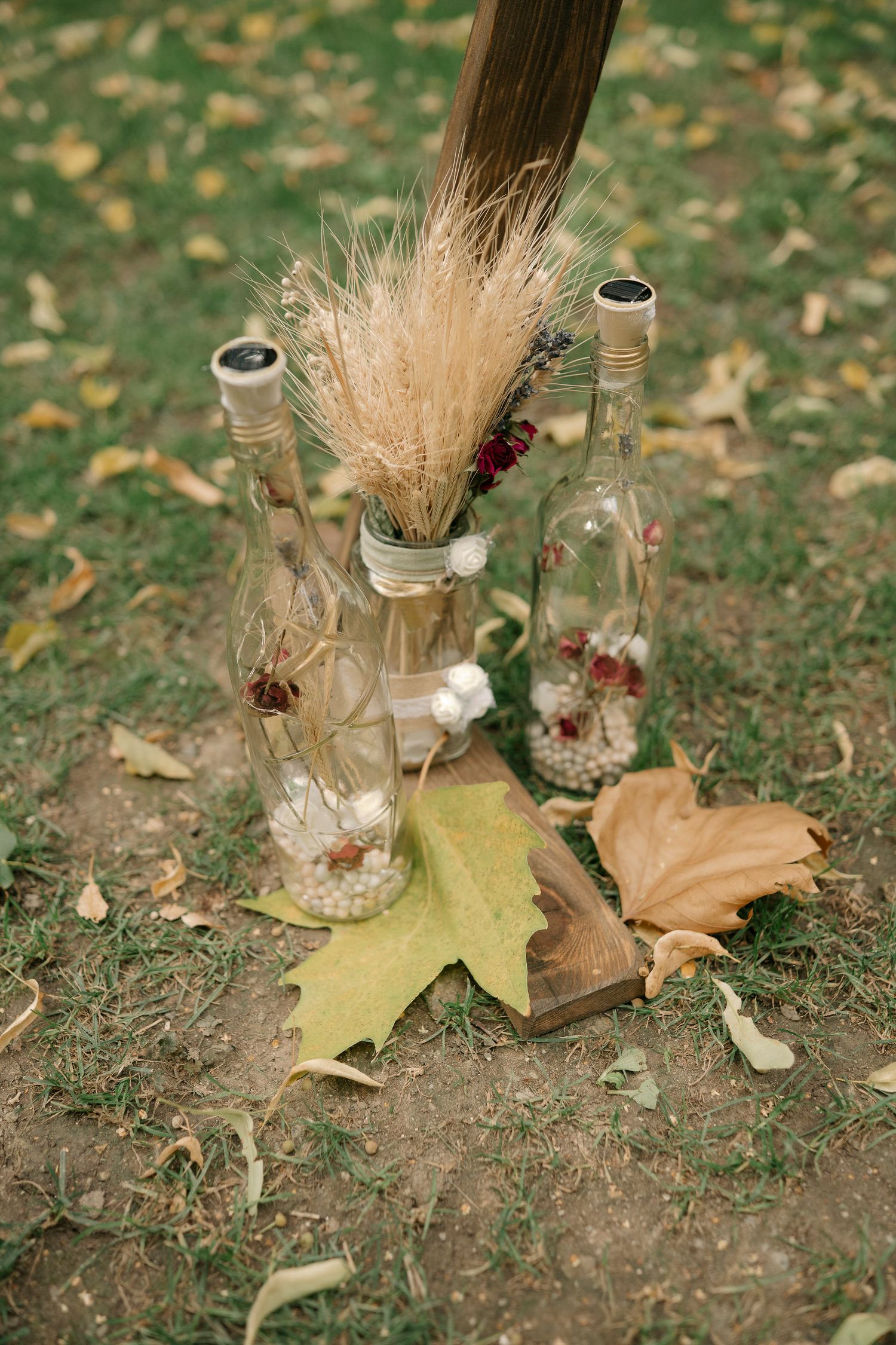Glass bottles with dried wheat stalks and autumn leaves create a rustic seasonal display on ground covered in fallen foliage.