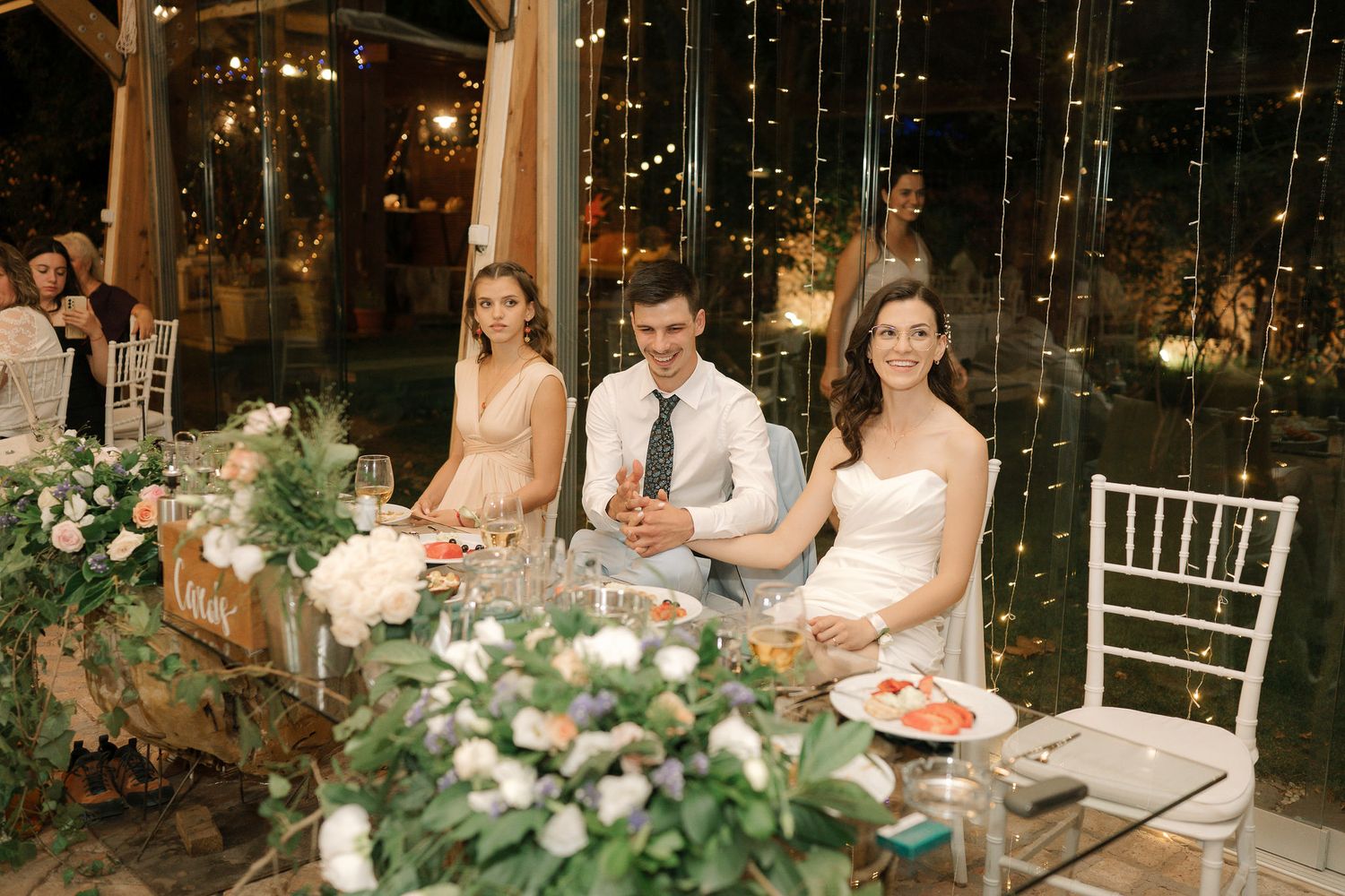 Wedding party seated at an elegantly decorated reception table with lush floral arrangements and twinkling lights overhead.