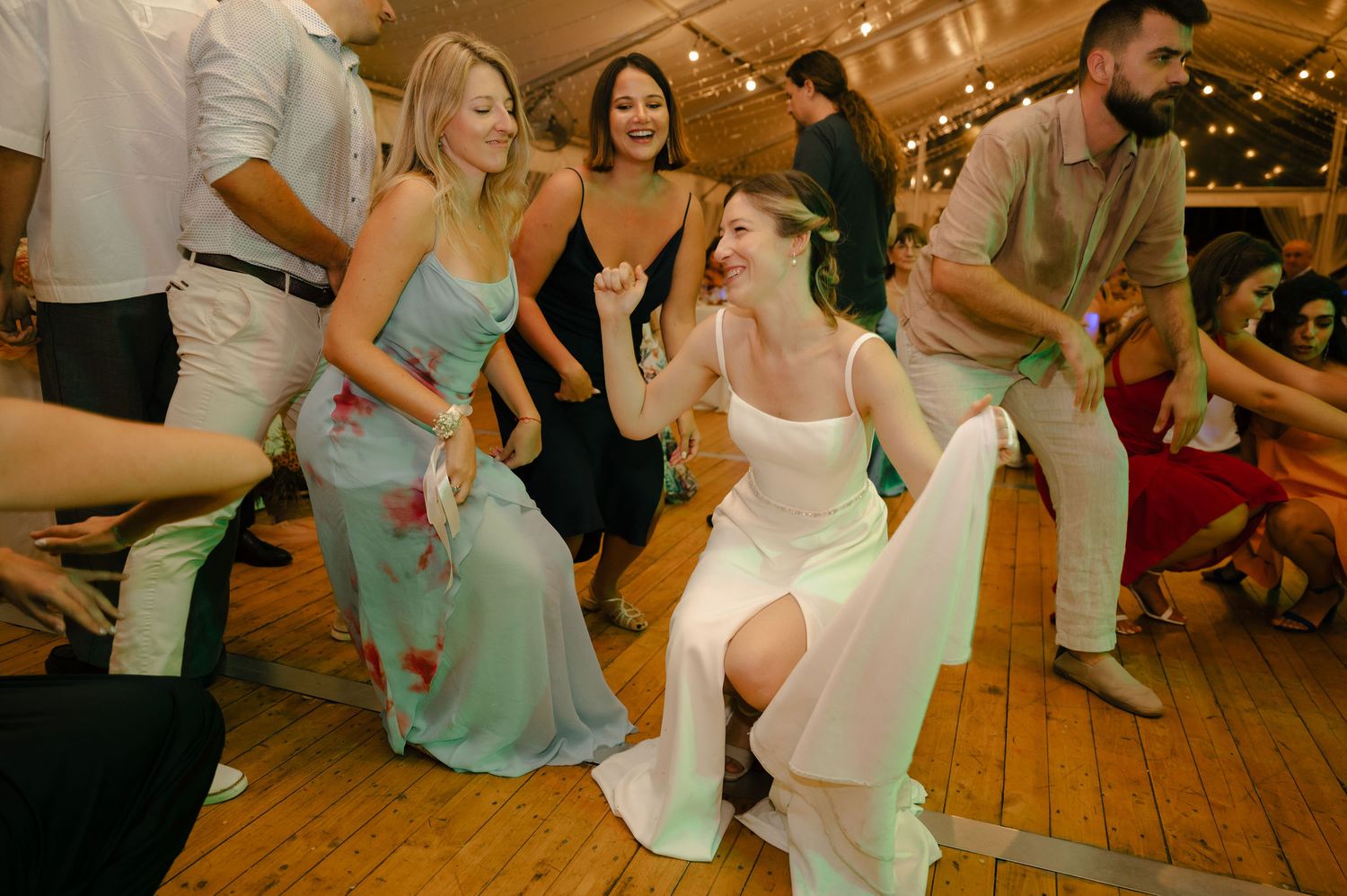 Guests dancing and celebrating at a wedding reception on a wooden dance floor with decorative lighting overhead.