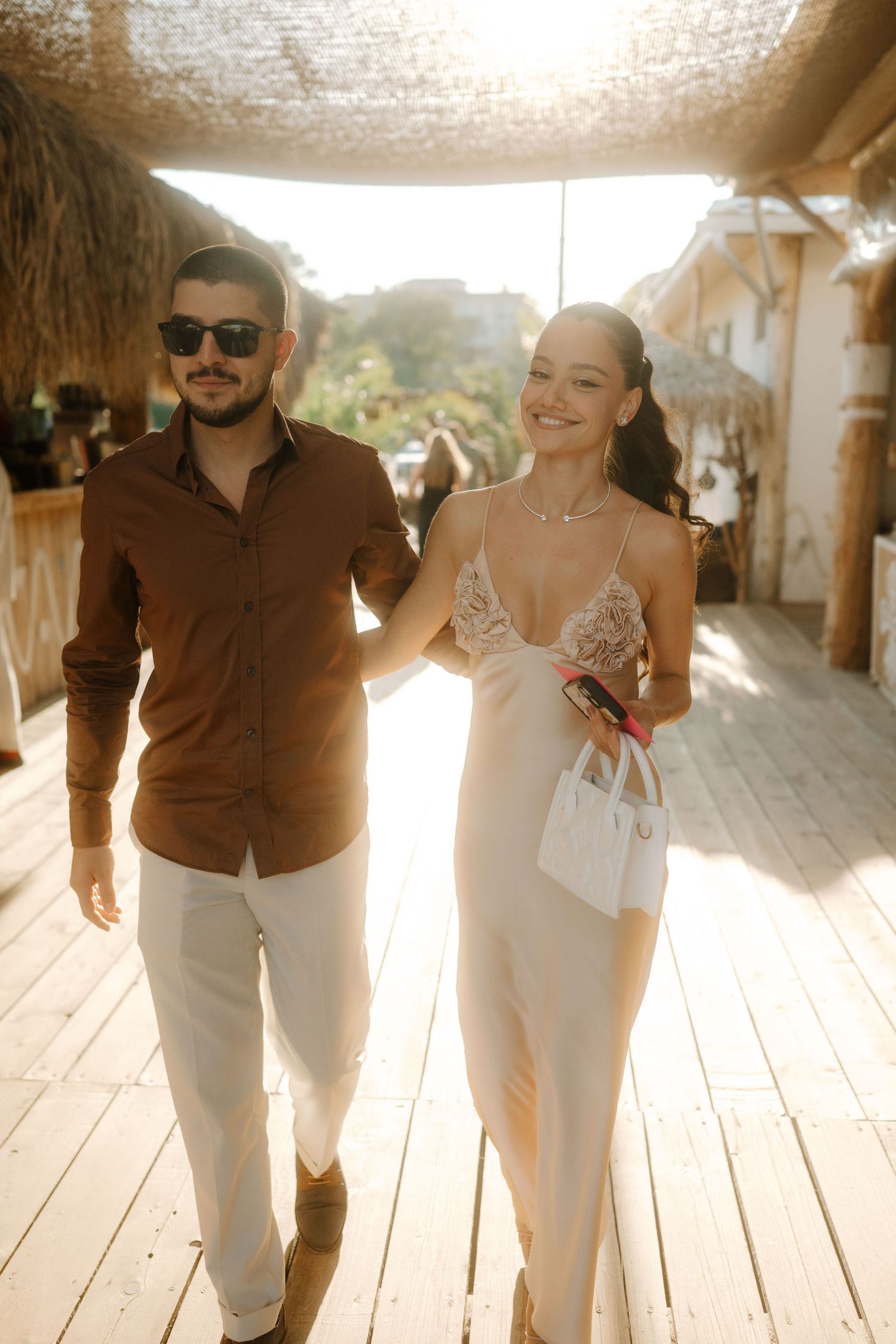 A couple walks together on a wooden deck at a tropical resort, wearing elegant summer attire in warm sunlight.