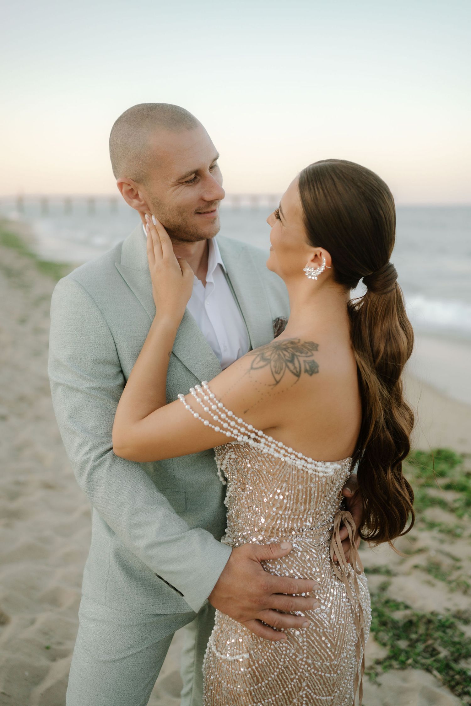 A romantic couple embraces on the beach at sunset, with her wearing a sparkly gown and him in a light suit.