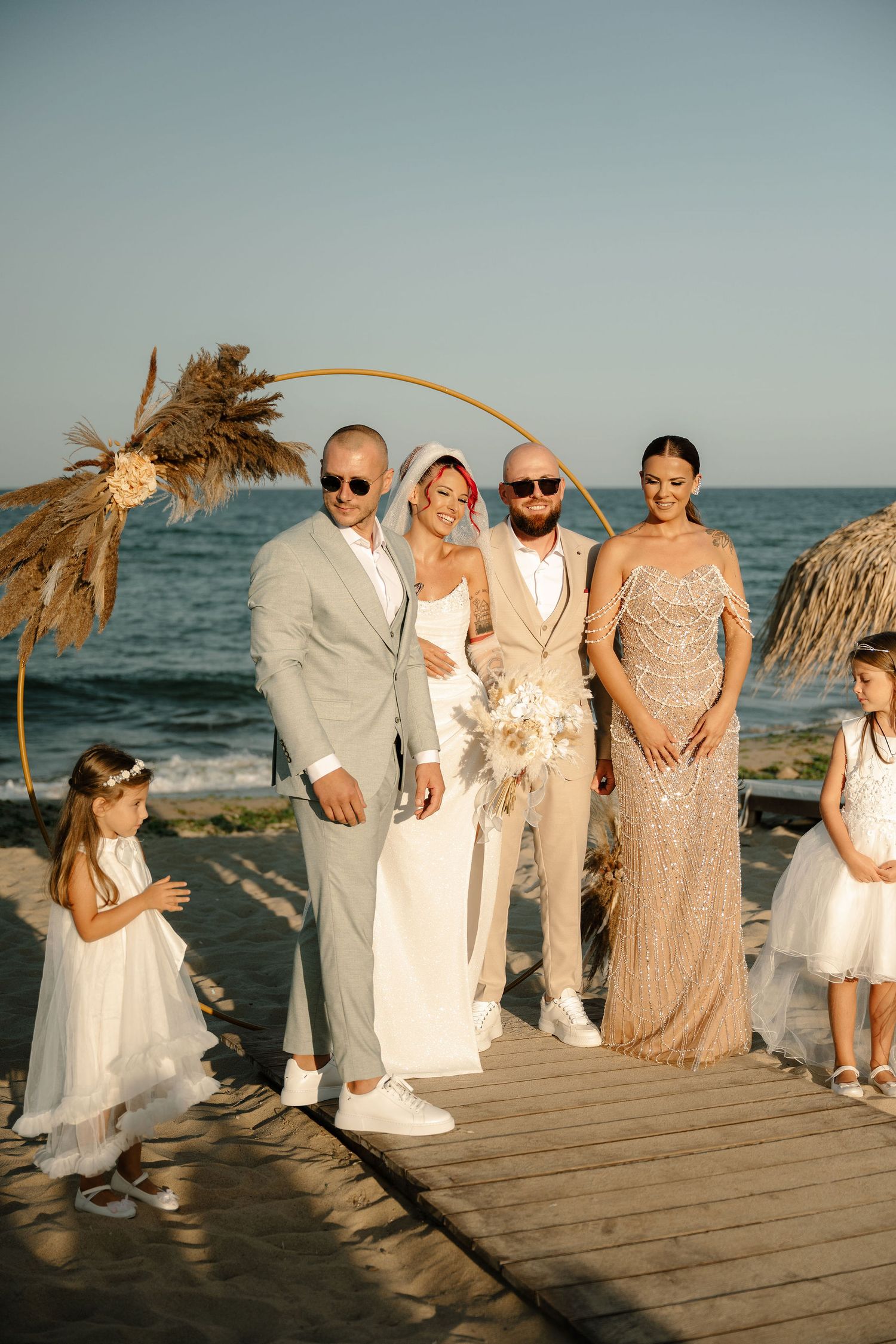 Wedding party dressed in white and beige poses on a wooden boardwalk by the ocean at sunset.