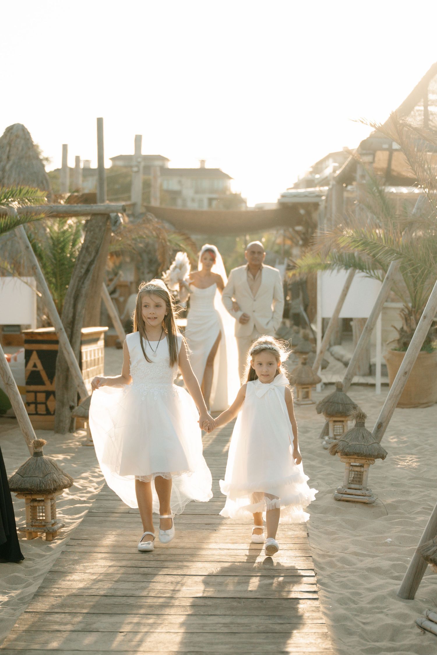 Young flower girls in white dresses walk down a beach boardwalk during a sunset wedding ceremony.