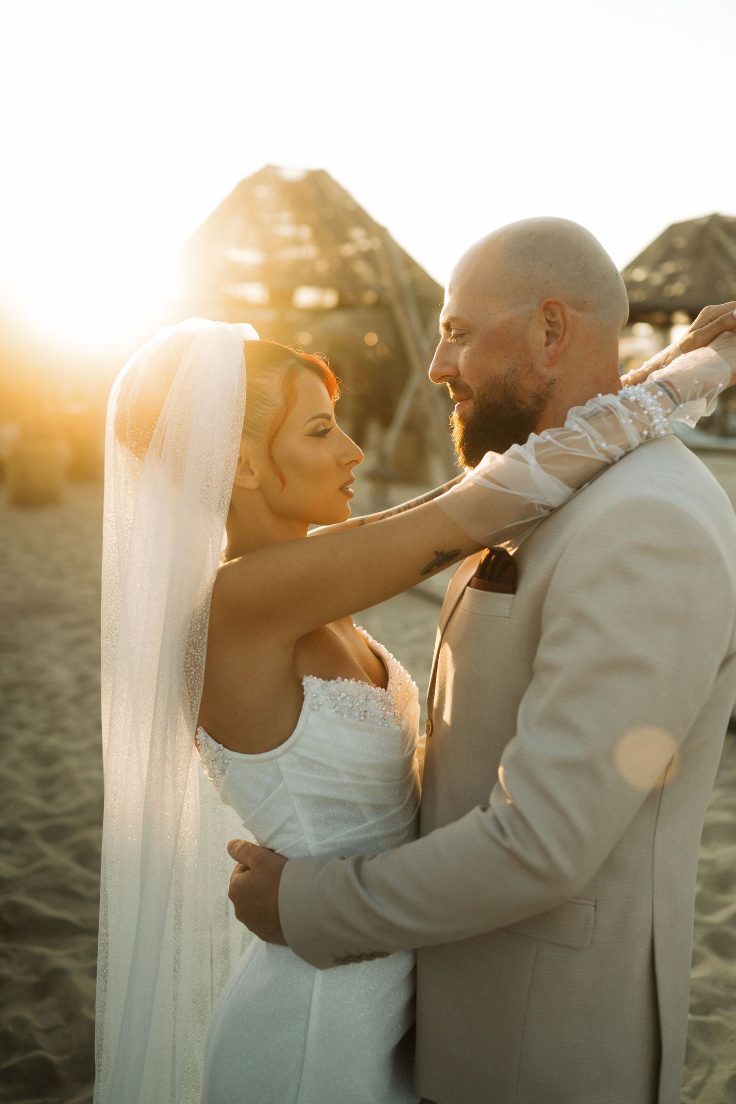 A romantic couple shares intimate moments at sunset on the beach, with the bride wearing a flowing white veil.