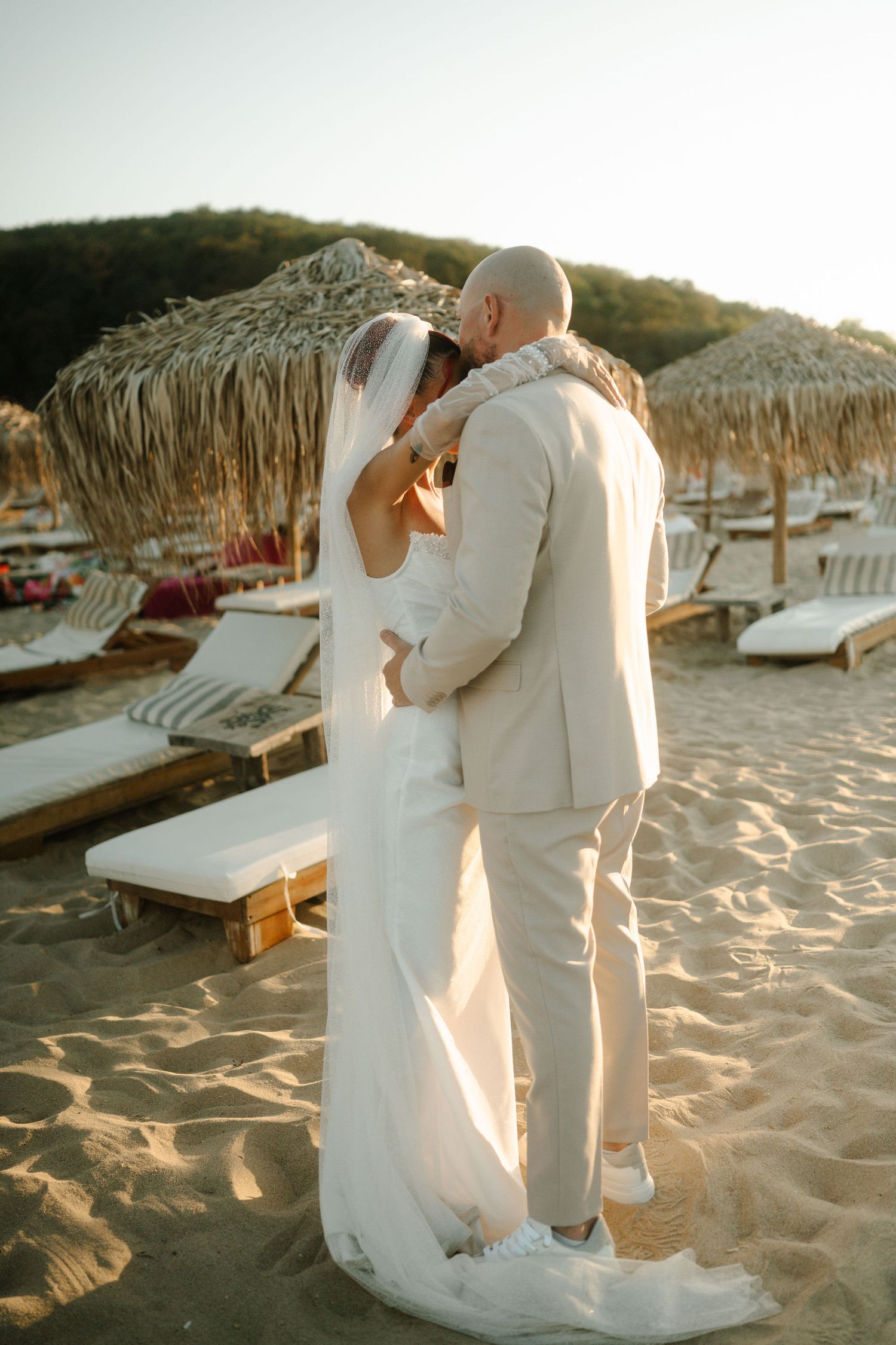 A romantic couple in white attire embraces on a sandy beach with thatched umbrellas and lounge chairs at sunset.