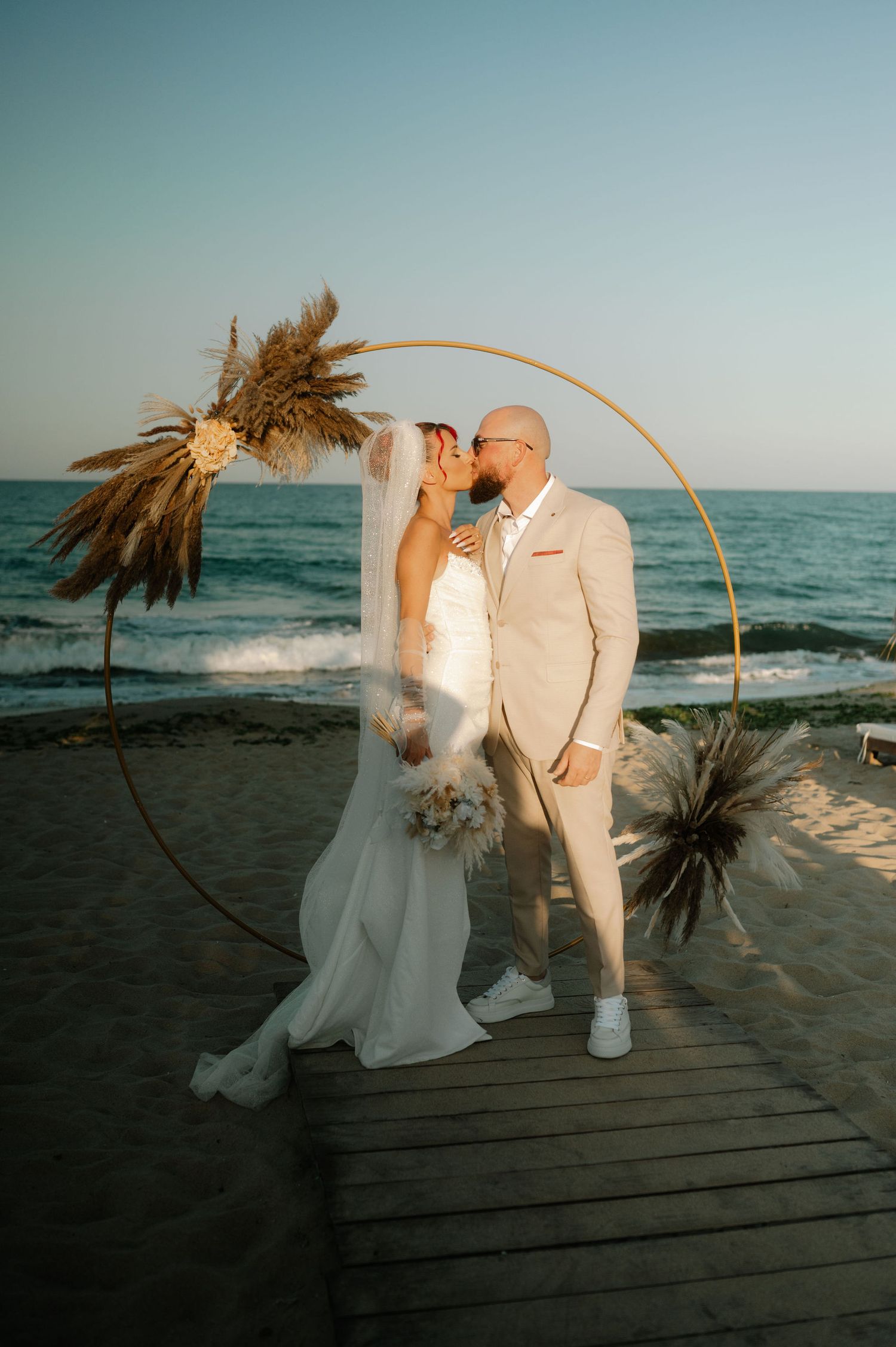 A couple shares a romantic moment on a beach boardwalk at sunset beneath a curved palm frond arch.