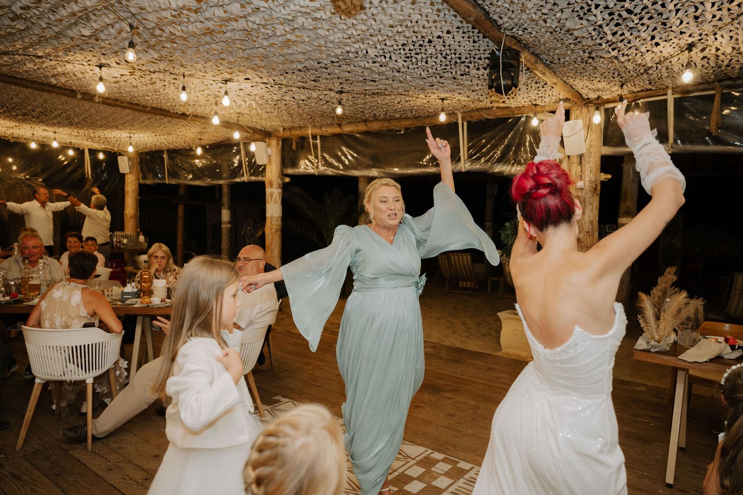 Wedding guests dance and celebrate under string lights in a rustic wooden venue during a reception.