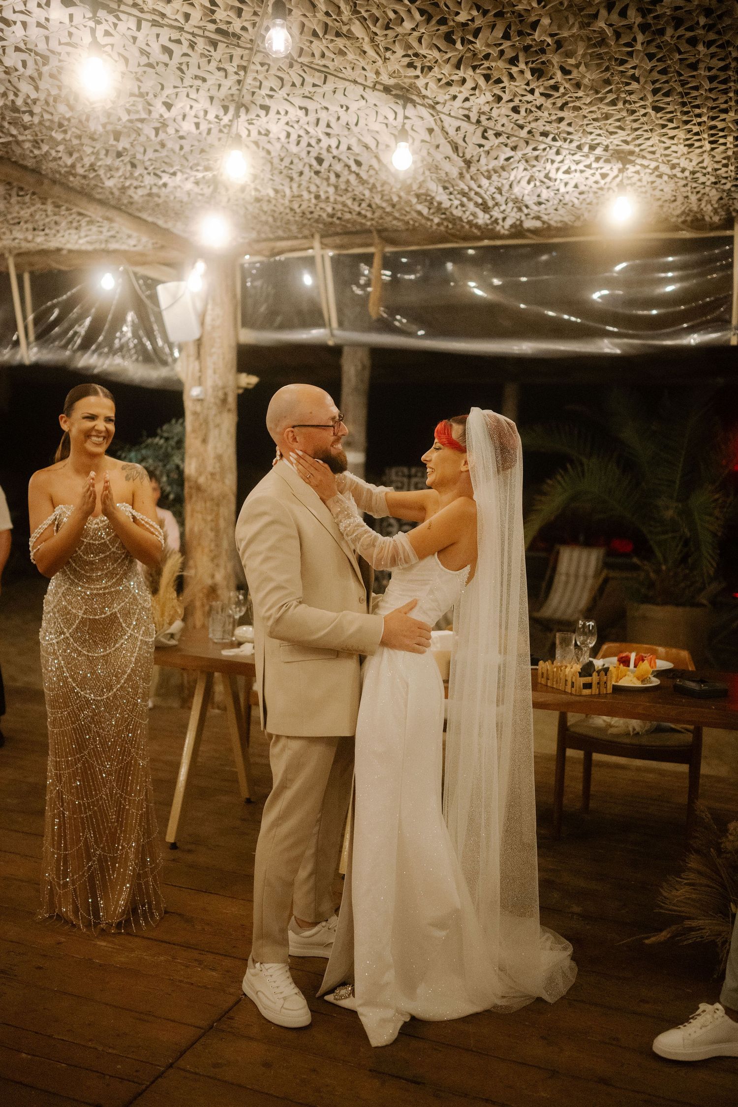 Wedding party celebrates on dance floor with joyful expressions under decorative ceiling lights at evening reception.