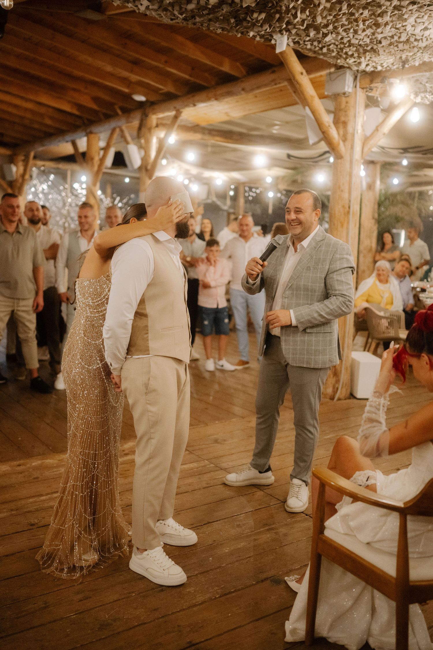 Wedding guests dance together on a rustic wooden dance floor in a barn venue illuminated by warm string lights.