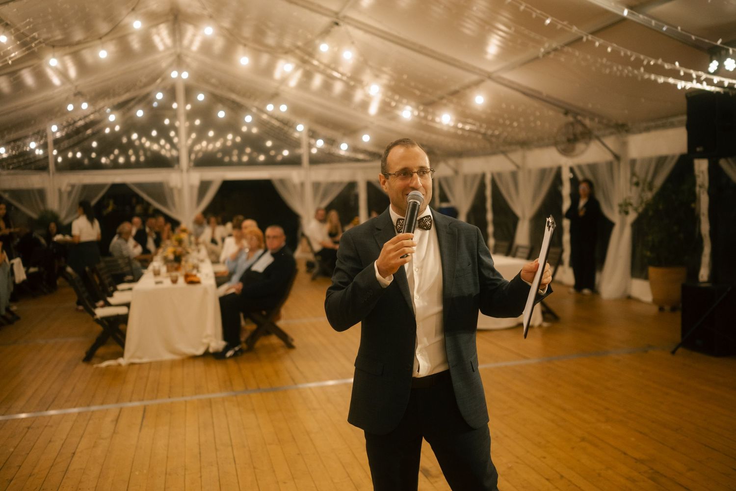 A person in a black tuxedo speaks into a microphone at an elegant wedding reception under a tent with twinkling lights.