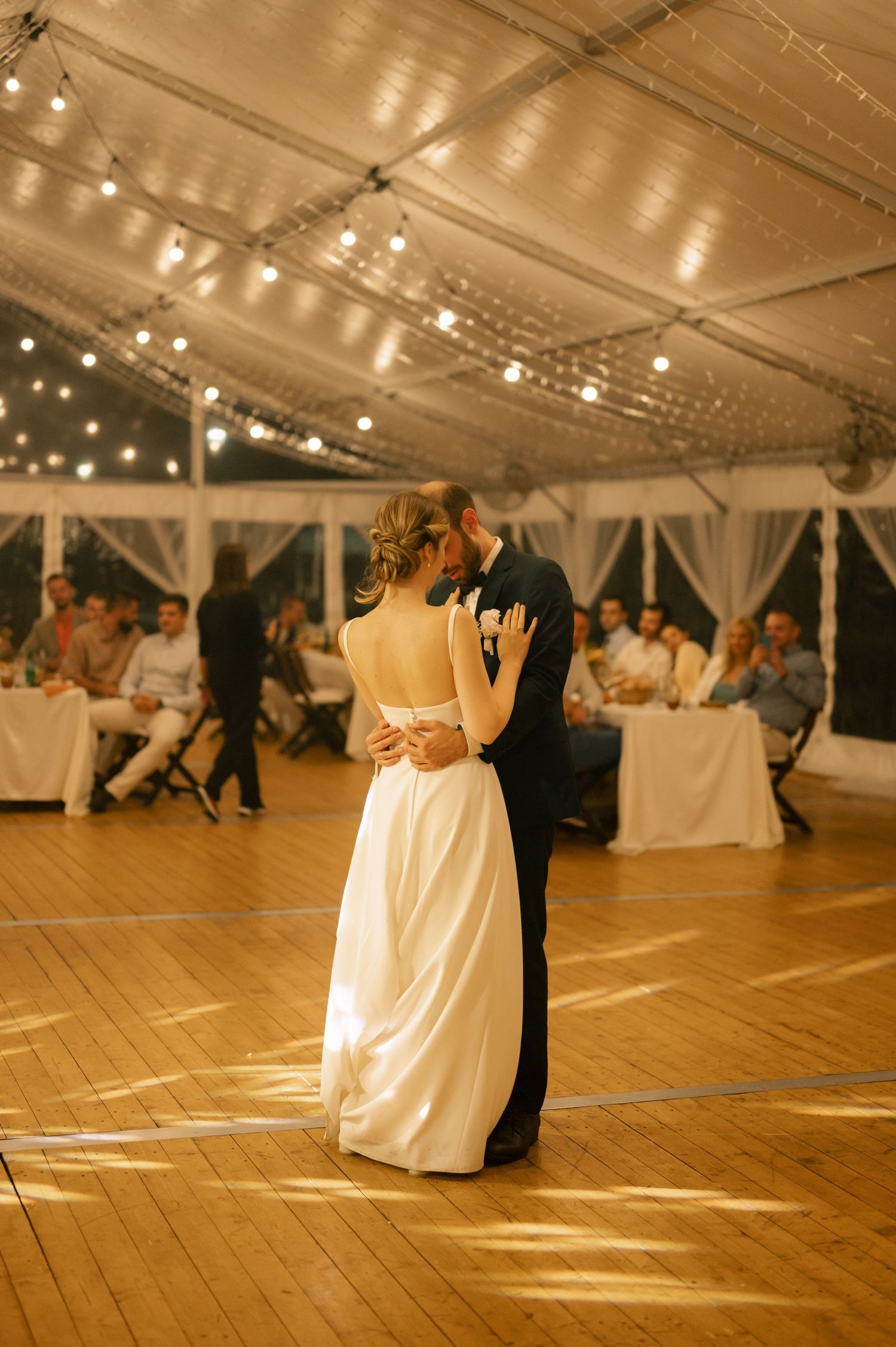 A couple dances on a wooden floor under a white tent with string lights at an elegant wedding reception.