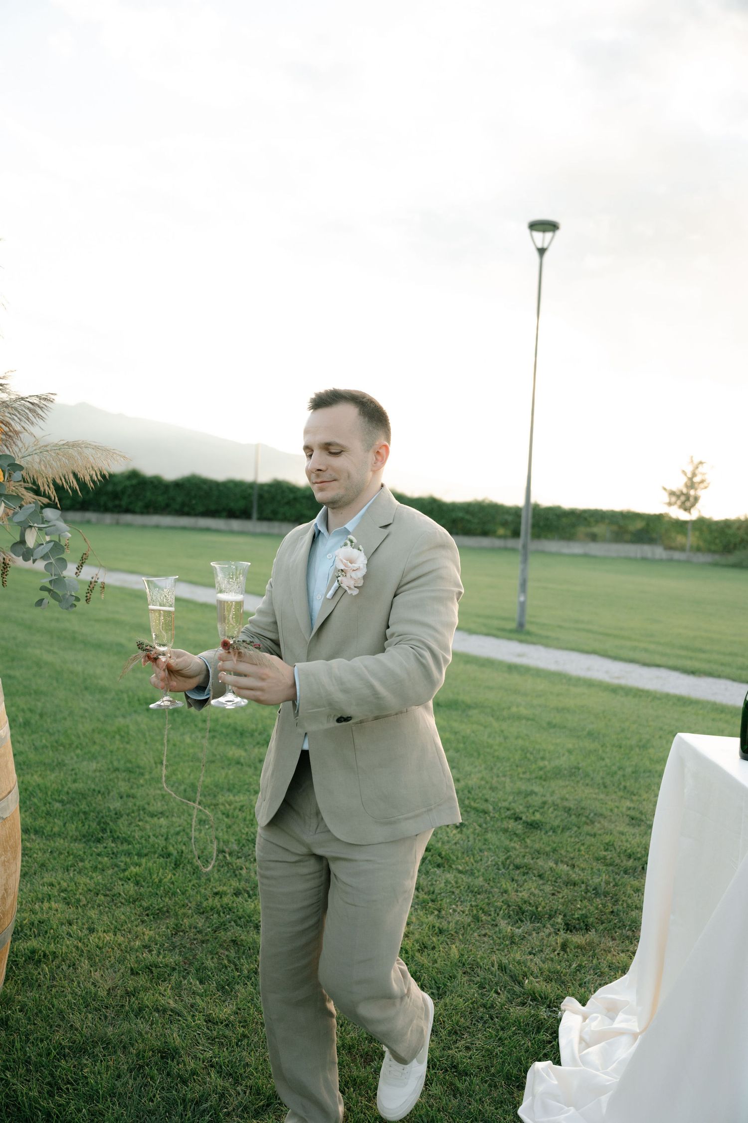 Wedding party member in a light gray suit checks their phone while standing on a grassy lawn at an outdoor event.