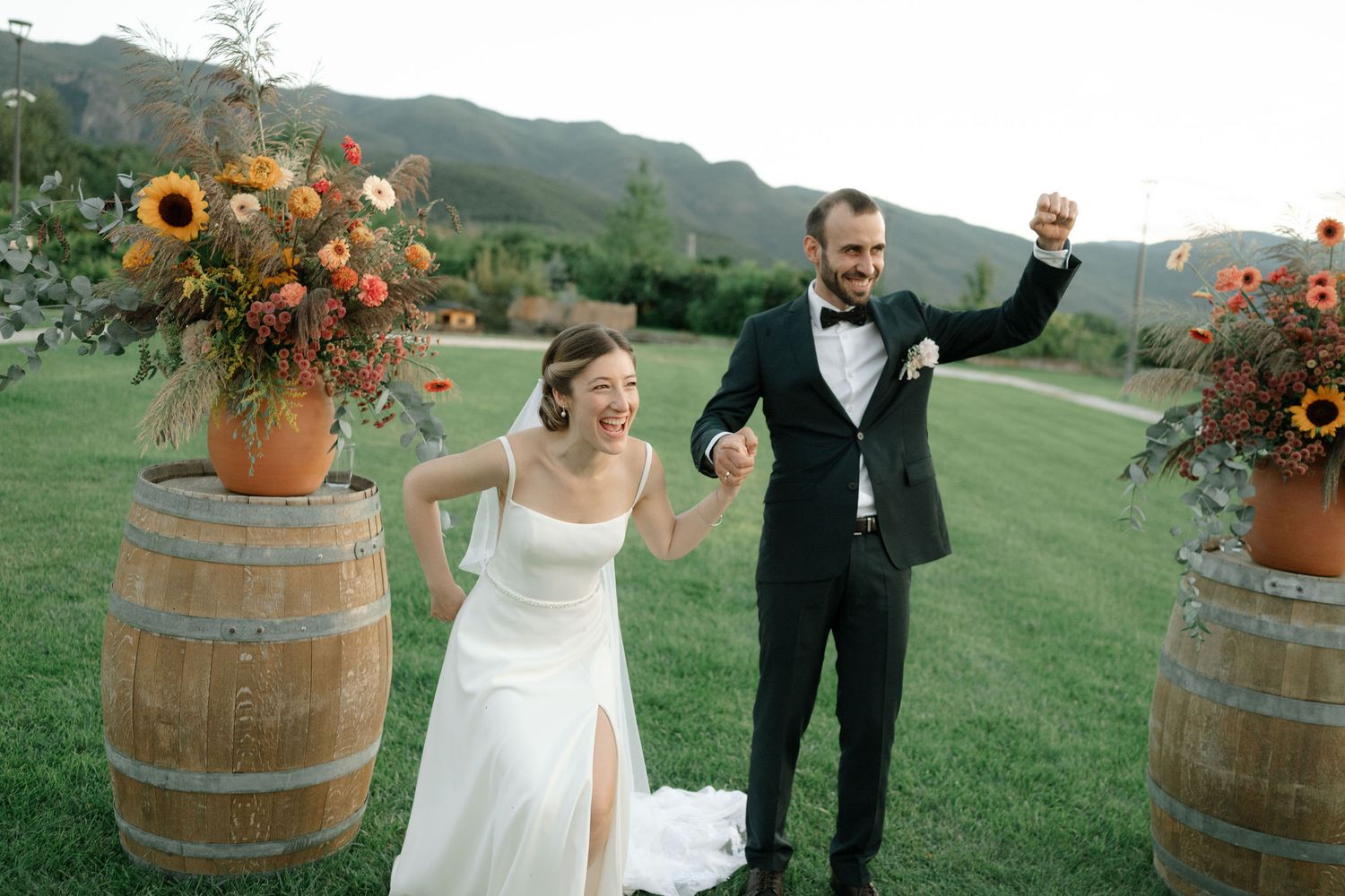 A joyful couple celebrates on a lawn with rustic wine barrel decor and vibrant sunflower arrangements in the background.