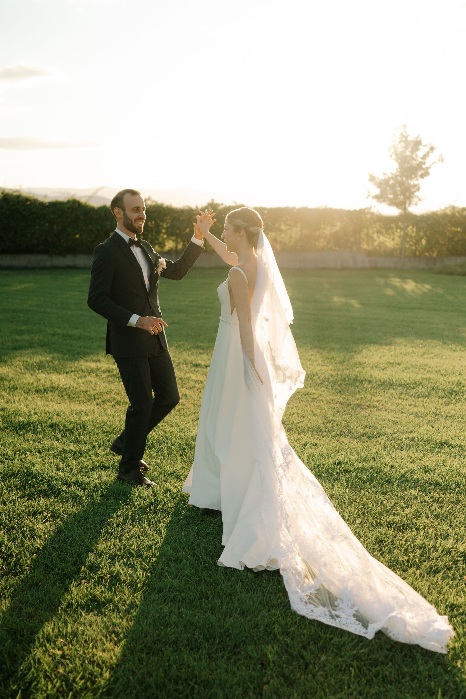 A couple in wedding attire dance together on a sunlit grassy field during golden hour.