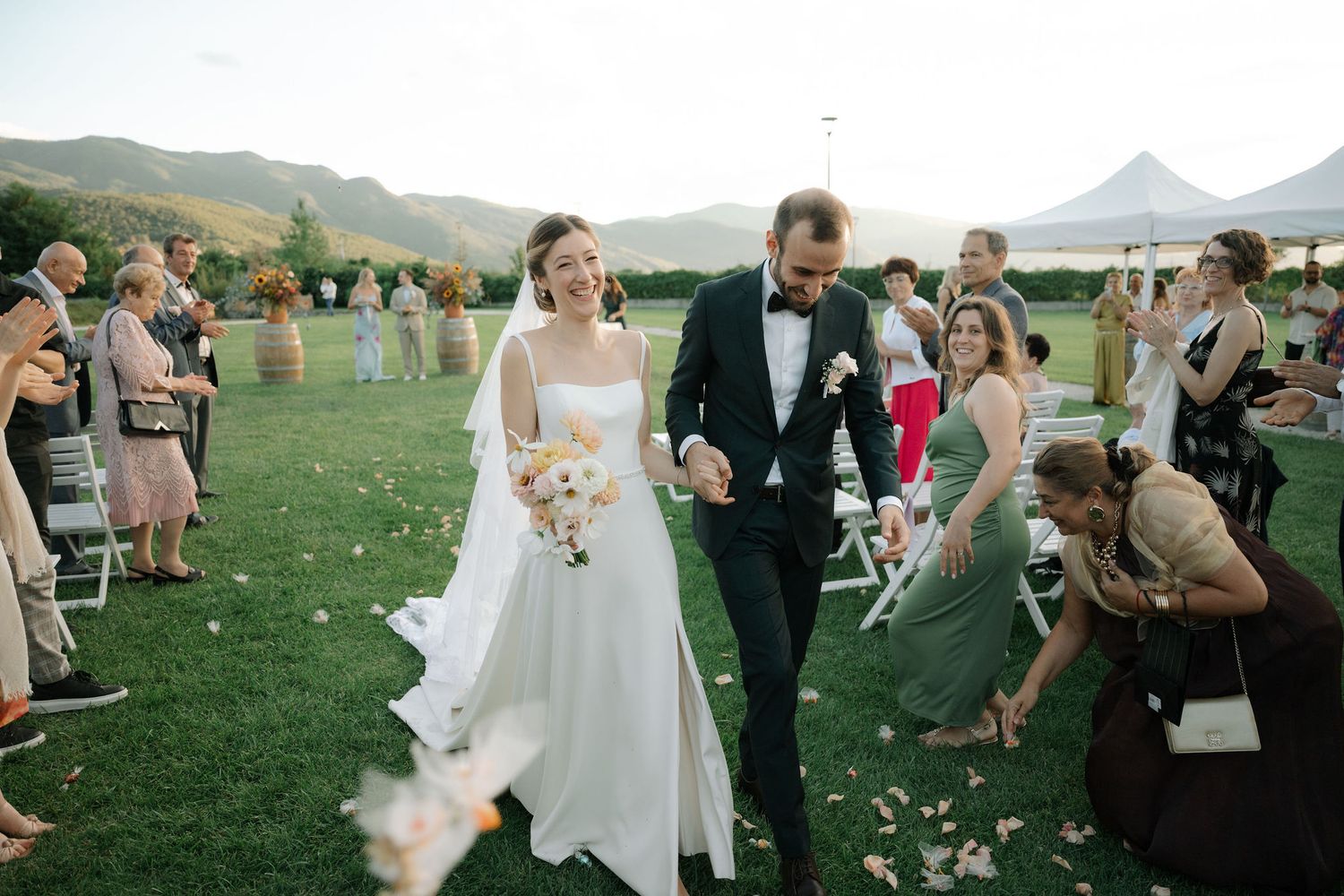Guests celebrate and toss flower petals at an outdoor wedding ceremony with mountain views in the background.